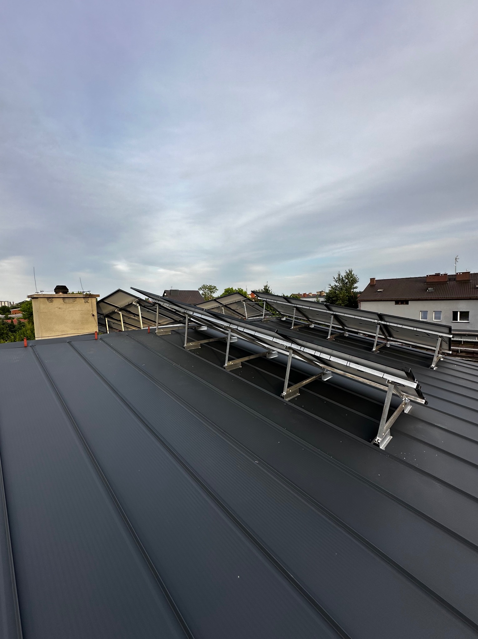 Instalacja paneli słonecznych na dachu z blachy trapezowej, widok z dołu pod kątem, w tle komin i zabudowa miejska pod pochmurnym niebem.
