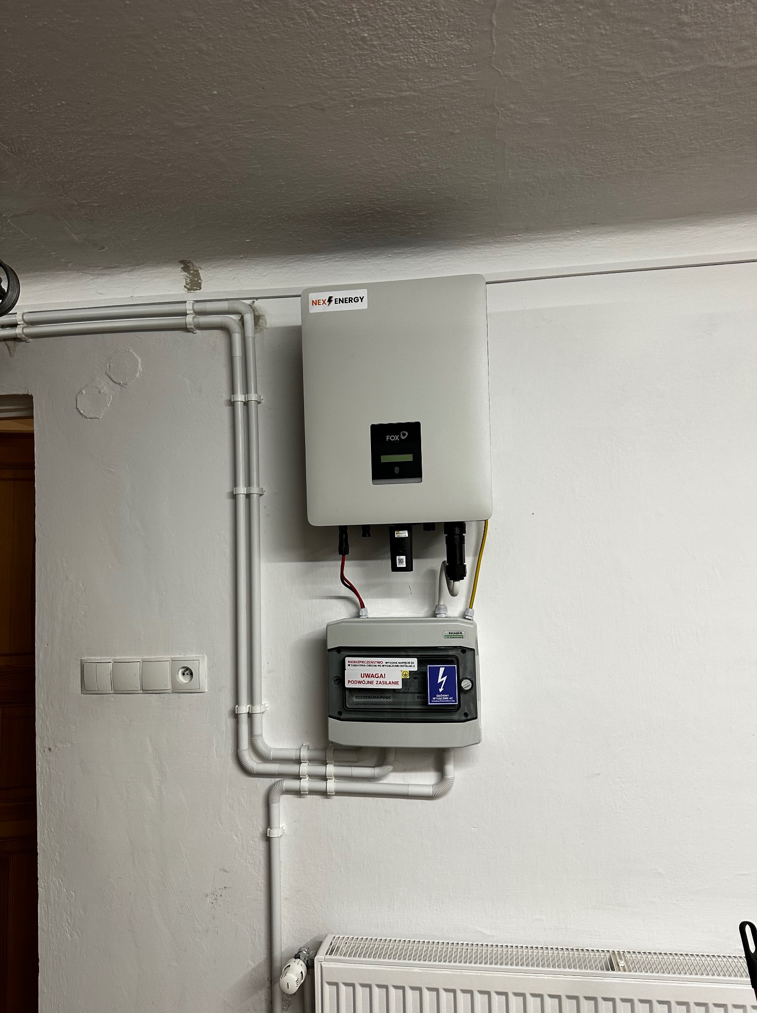 Zamontowany inwerter NEX ENERGY FOXO wraz z zabezpieczeniami DC wewnątrz pomieszczenia, widoczne białe rury instalacyjne i grzejnik.