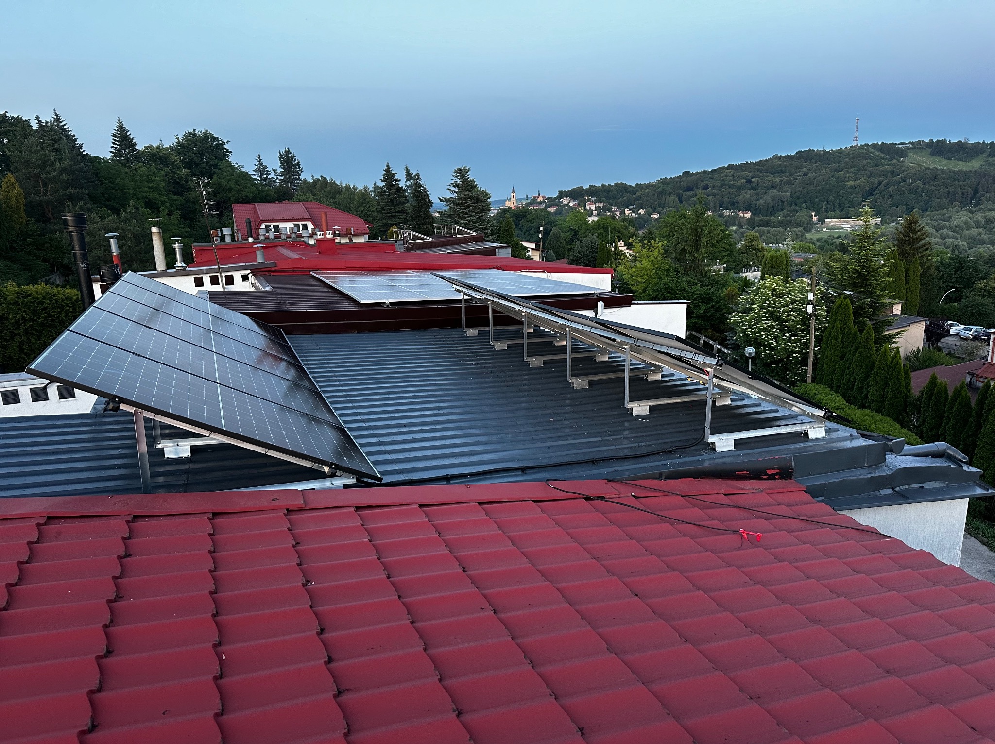 Instalacja fotowoltaiczna na dachu z blachy trapezowej, widok z góry na tle zabudowań i zalesionych wzgórz w oddali.