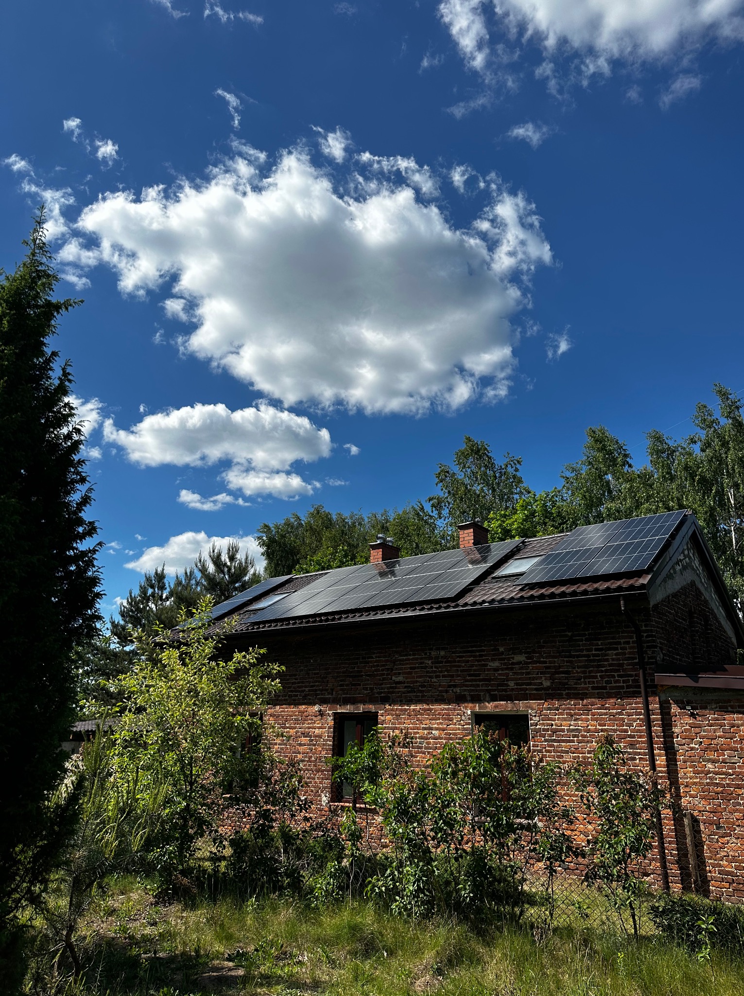 Ceglany budynek z panelami słonecznymi na dachu, widoczne kominy, błękitne niebo z białymi obłokami w tle.