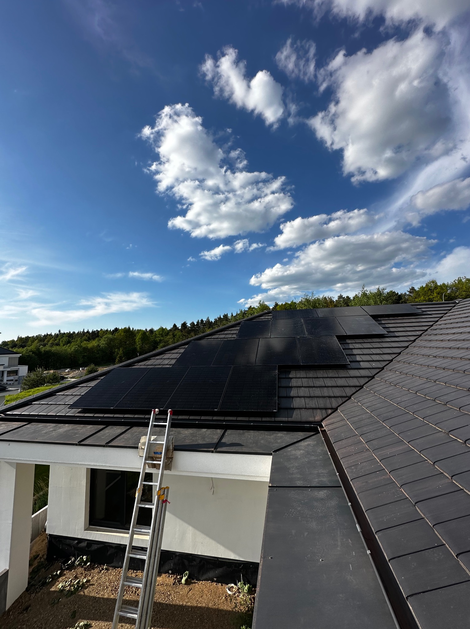 Montaż paneli słonecznych na dachu domu jednorodzinnego z użyciem drabiny, widok z góry na częściowo ukończoną instalację w słoneczny dzień.