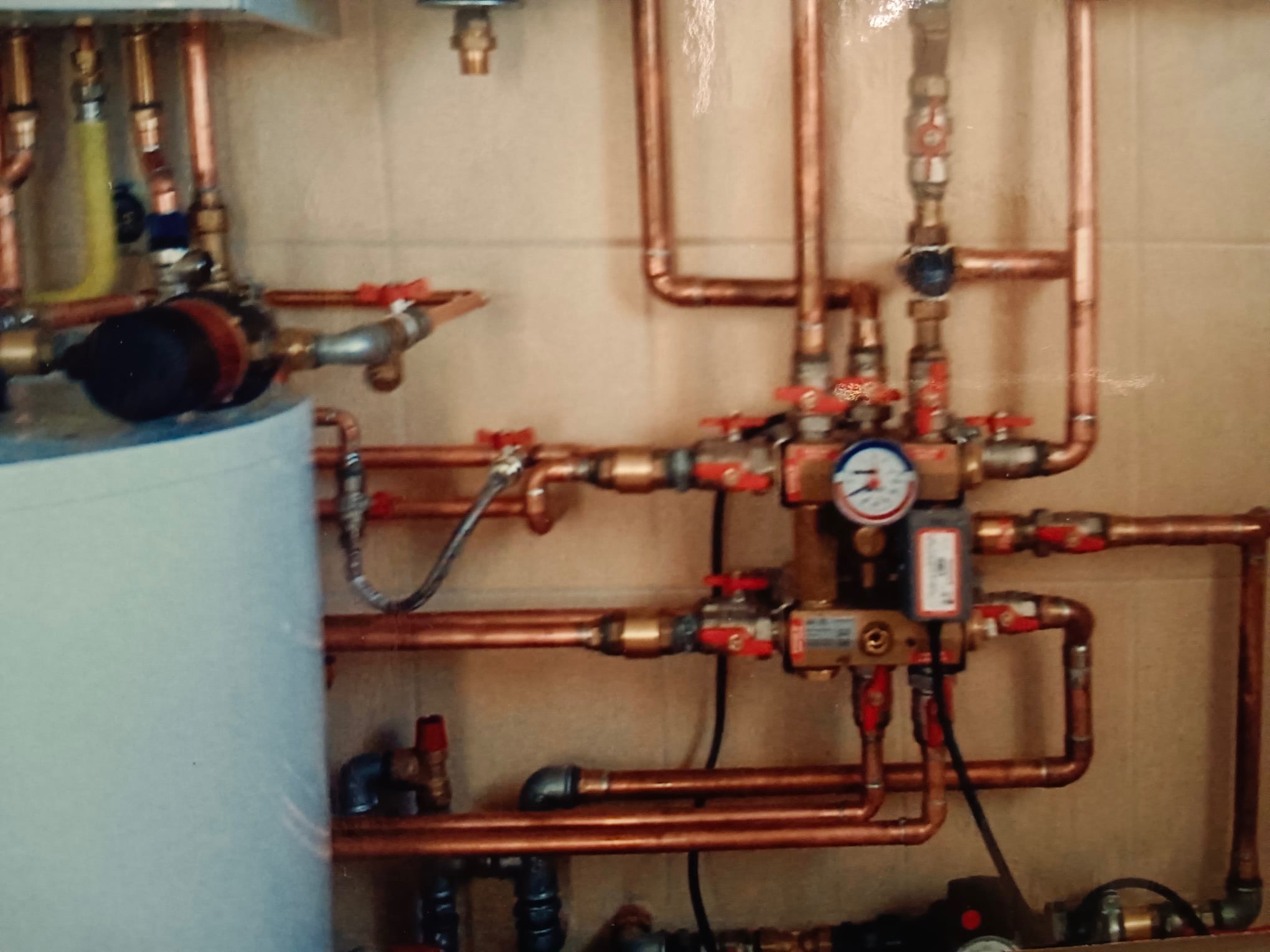 Złożony system rur miedzianych z zaworami i manometrem, część instalacji grzewczej w pomieszczeniu z beżowymi płytkami.