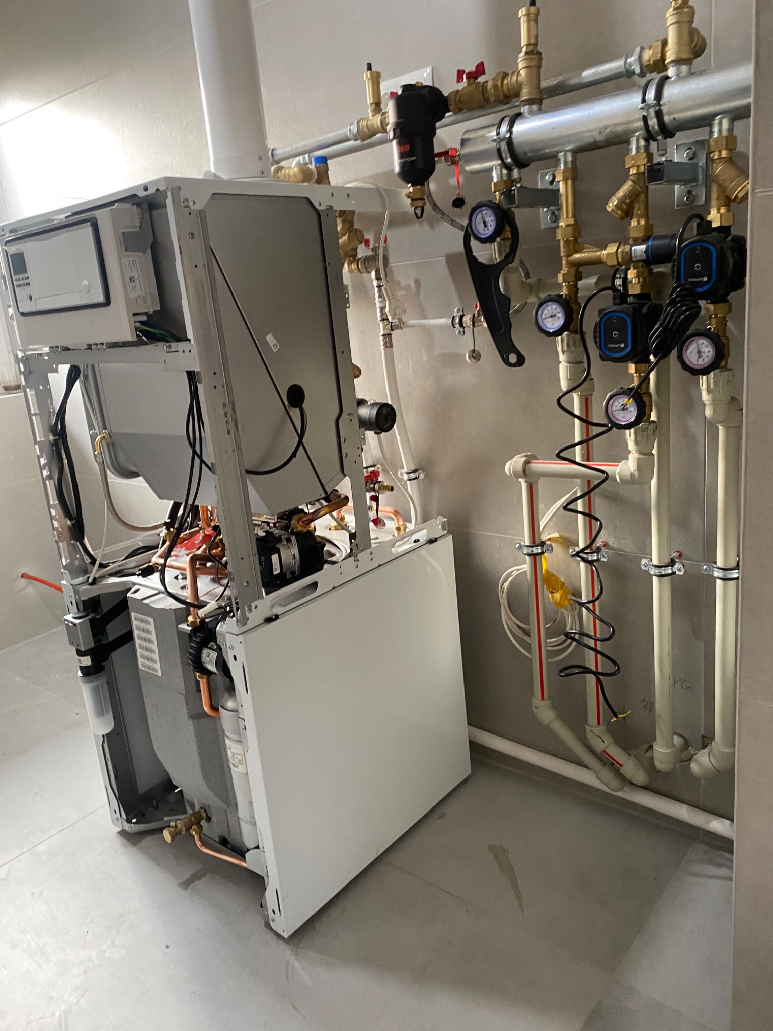 Wnętrze kotłowni: biały piec gazowy w trakcie montażu, widoczne podłączenia rur miedzianych i plastikowych, manometry, pompy obiegowe oraz filtry na ścianie wyłożonej szarą płytką.
