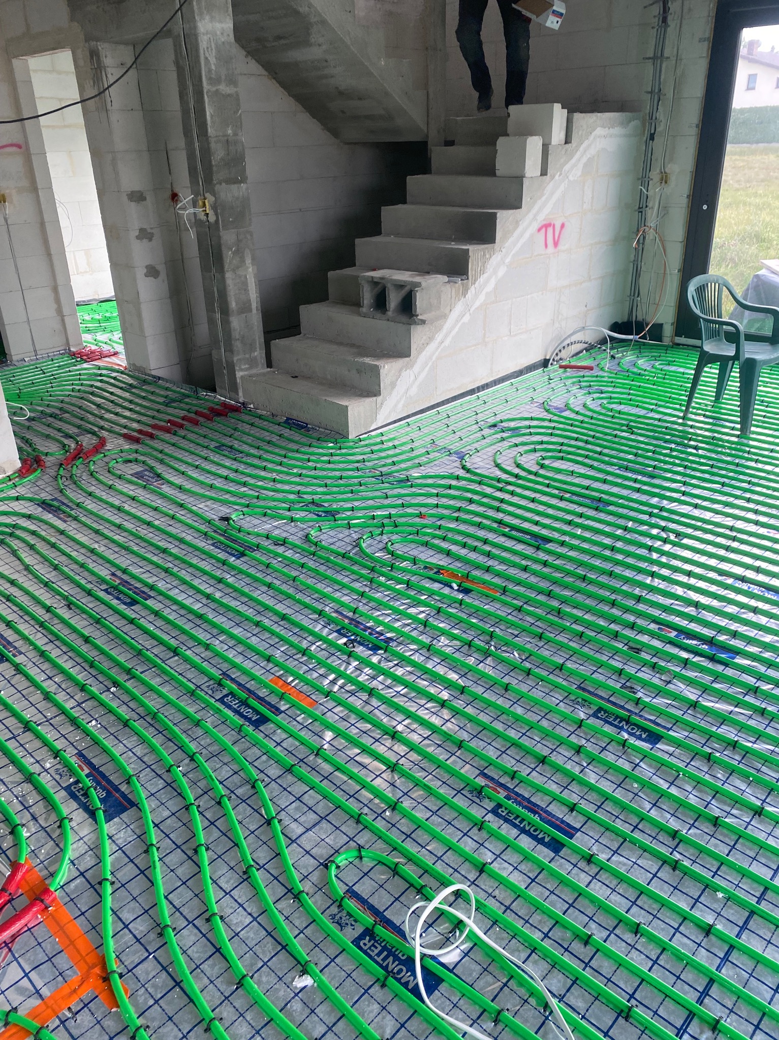 Wnętrze w stanie surowym z rozłożoną instalacją ogrzewania podłogowego: zielone rurki na siatce, betonowe schody w tle, osoba wchodząca po schodach.