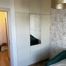 M.B. Meblox Beata Kosińska - Biała szafa z lustrzanym frontem, widok z boku na tle wzorzystej tapety i złotego abażuru, z widocznym fragmentem zielonego łóżka w folii.