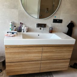 M.B. Meblox Beata Kosińska - Nowoczesna szafka łazienkowa z drewnianymi frontami szuflad, biała umywalka nablatowa, okrągłe lustro z odbiciem kabiny prysznicowej, akcesoria łazienkowe na blacie, widoczny wiertarko-wkrętak...