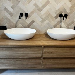 M.B. Meblox Beata Kosińska - Łazienka z dwiema owalnymi umywalkami nablatowymi na drewnianej szafce z szufladami, czarne baterie ścienne i beżowe płytki ułożone w jodełkę.
