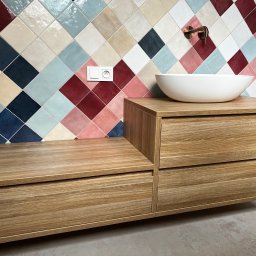 M.B. Meblox Beata Kosińska - Drewniana szafka łazienkowa z dwiema szufladami i białą umywalką nablatową, na tle ściany z kolorowych, romboidalnych płytek ceramicznych i miedzianej baterii.
