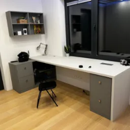 Nowoczesne biurko na wymiar z szarymi szafkami po bokach, biały blat, czarne krzesło składane, półka wisząca z dekoracjami, duże okno z czarną ramą, drewniana podłoga.