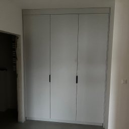 M.B. Meblox Beata Kosińska - Trzydrzwiowa szafa wnękowa z szarymi frontami i czarnymi, pionowymi uchwytami, widoczna w trakcie montażu, z surowym otworem drzwiowym w tle.