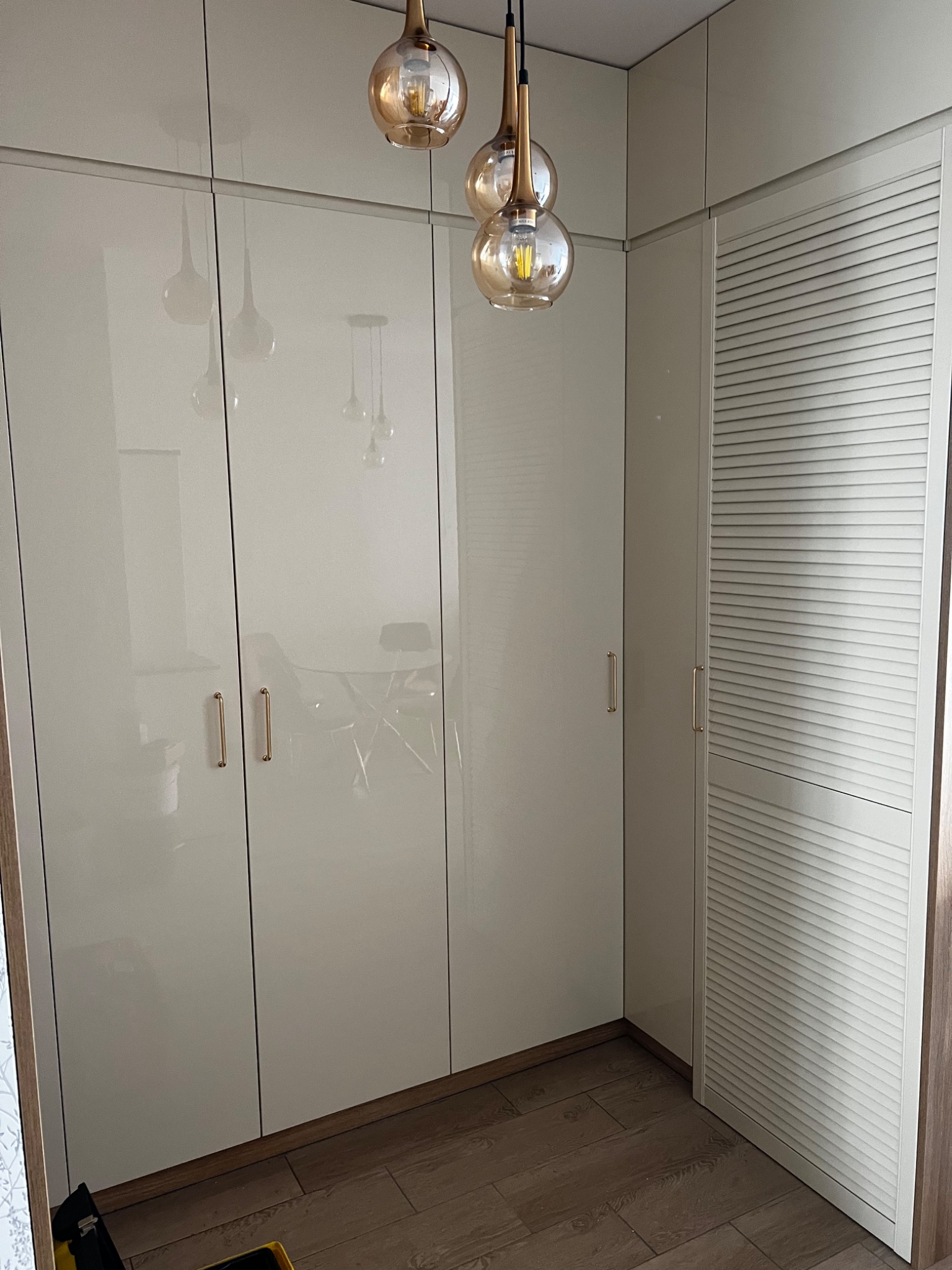 Biała, błyszcząca zabudowa meblowa z szafkami i drzwiami żaluzjowymi, oświetlona wiszącymi lampami z bursztynowego szkła.