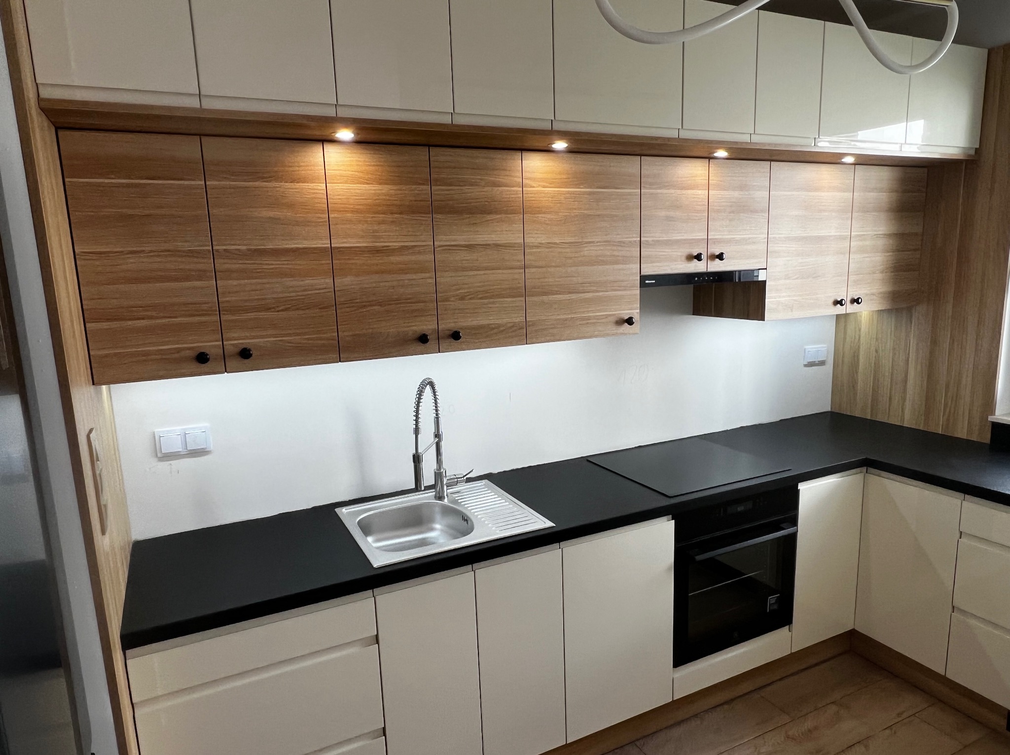 Nowoczesna kuchnia z drewnianymi szafkami górnymi i beżowymi dolnymi, czarny blat, stalowy zlew z baterią i płyta indukcyjna.