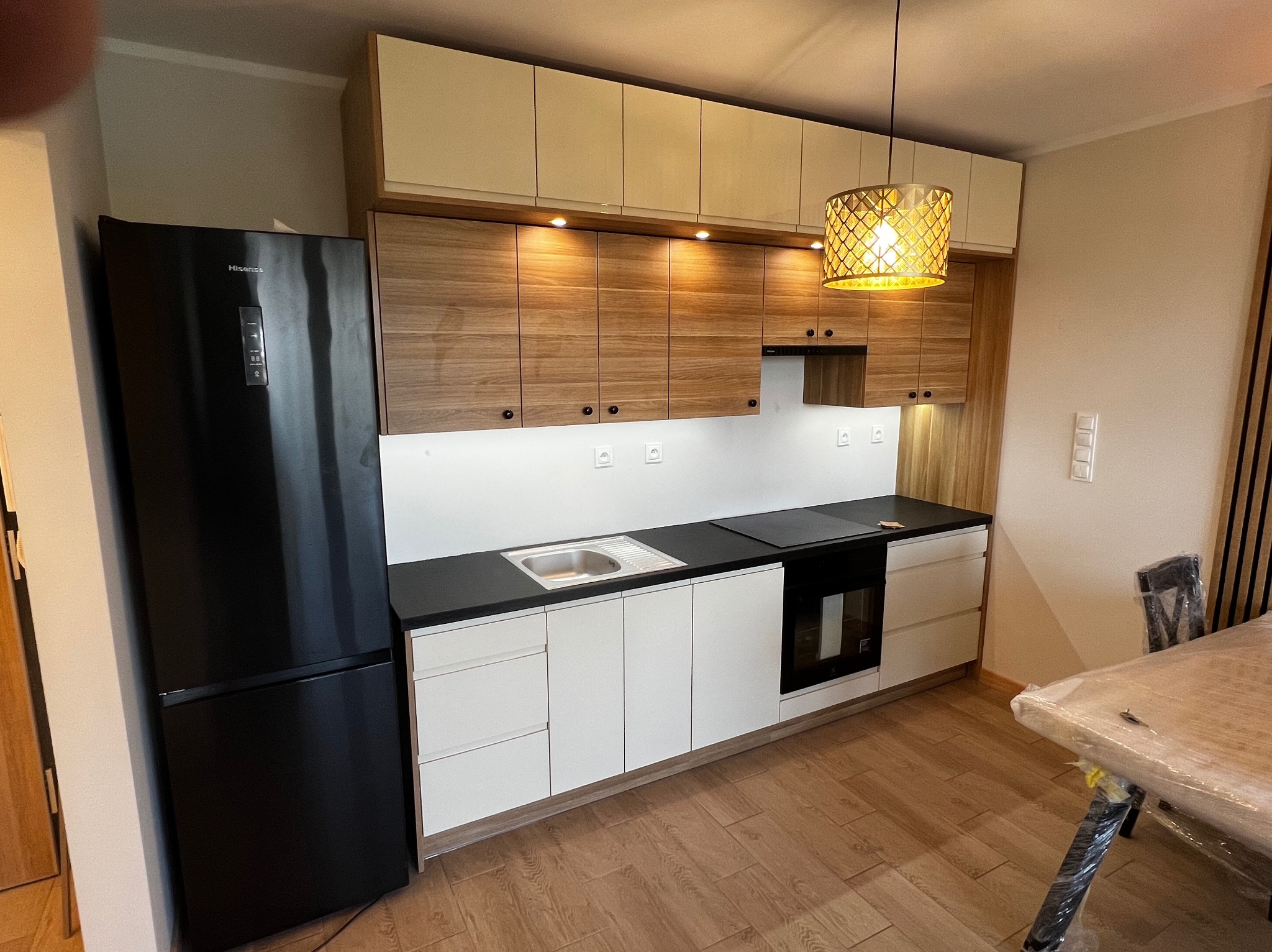 Nowoczesna kuchnia na wymiar z białymi frontami dolnymi, drewnopodobnymi górnymi szafkami i czarnym blatem, zintegrowana z czarną lodówką Hisense, oświetlona dekoracyjną lampą wiszącą.