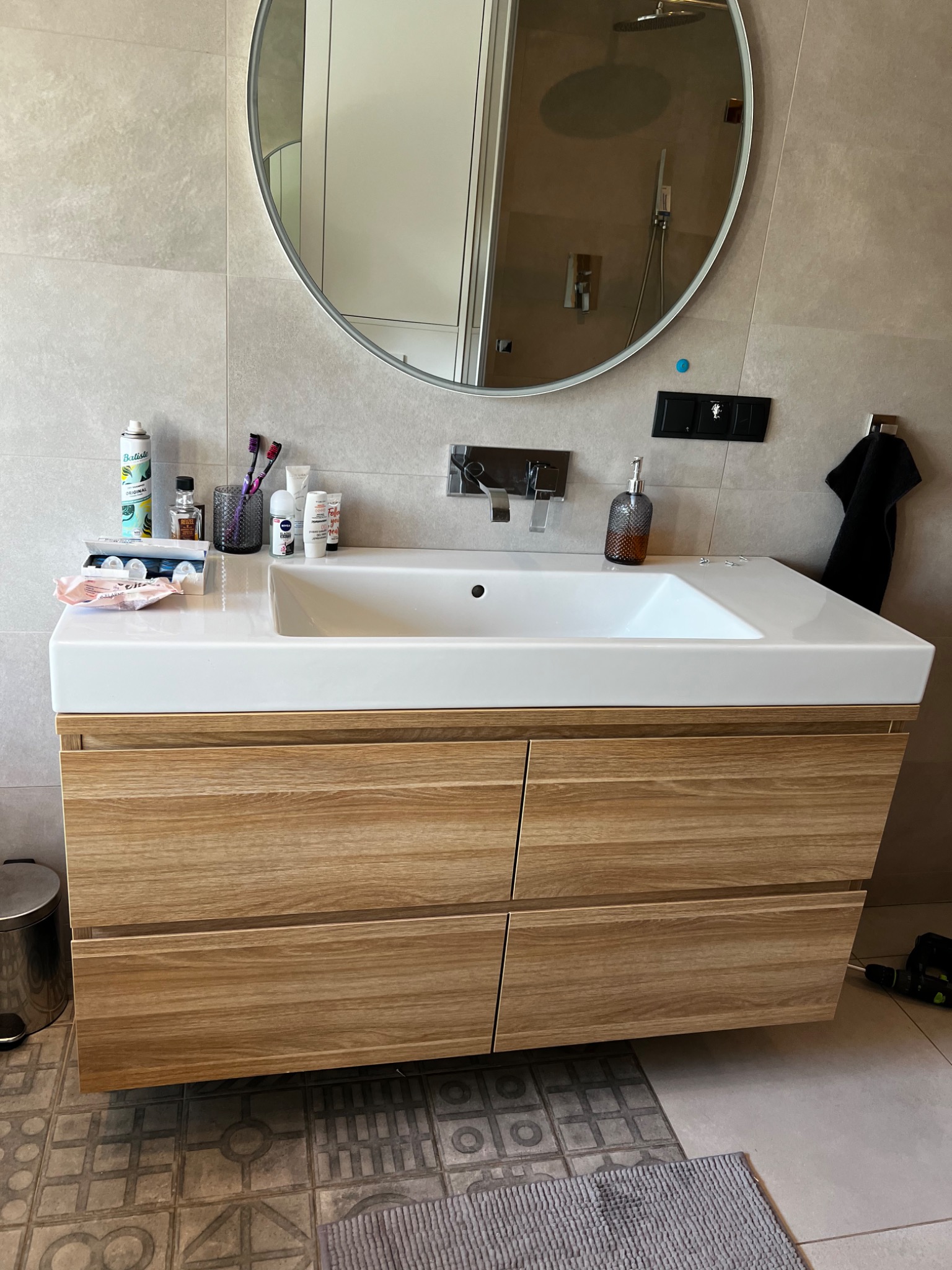 Nowoczesna szafka łazienkowa z drewnianymi frontami szuflad, biała umywalka nablatowa, okrągłe lustro z odbiciem kabiny prysznicowej, akcesoria łazienkowe na blacie, widoczny wiertarko-wkrętak...