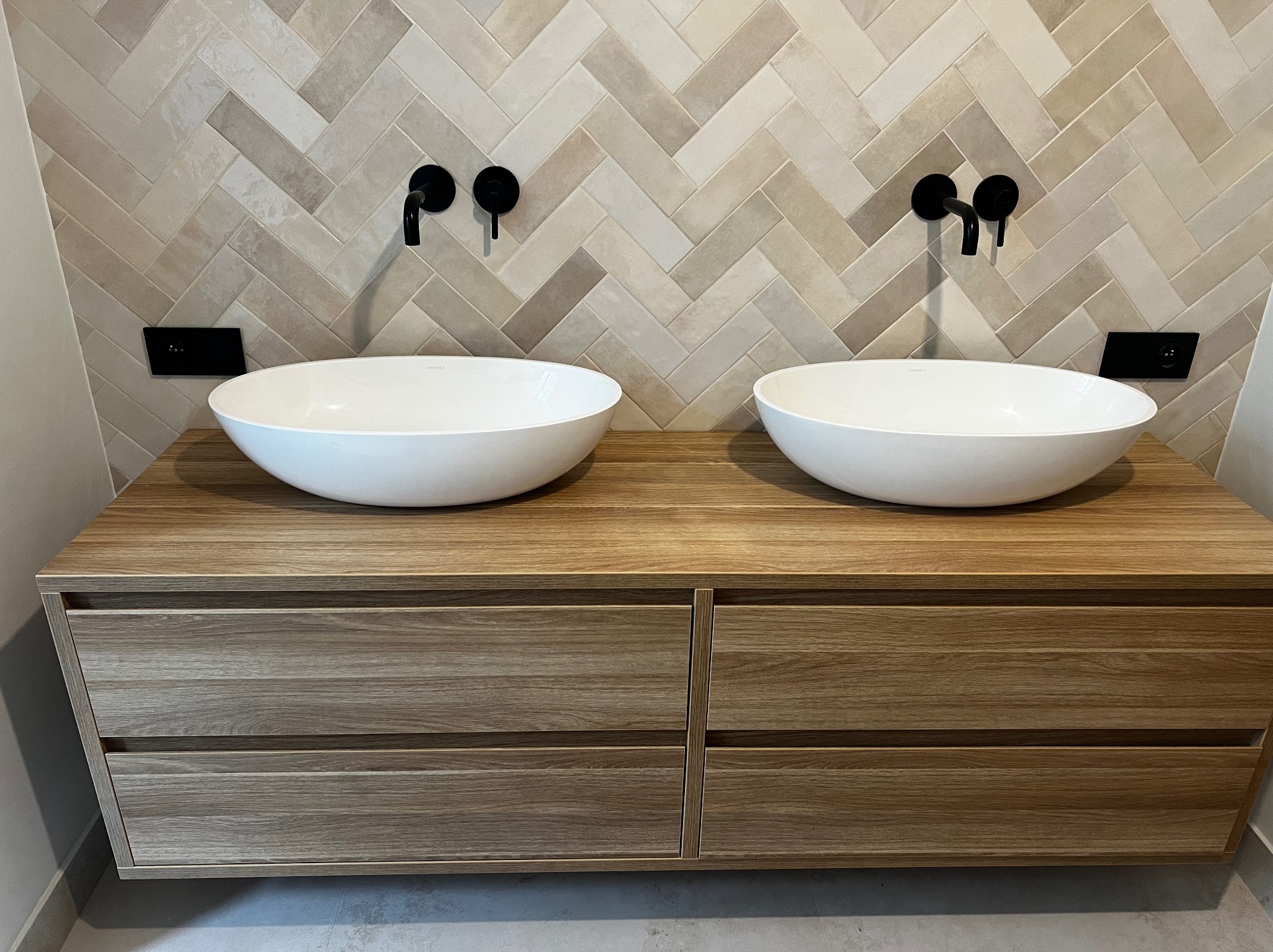 Łazienka z dwiema owalnymi umywalkami nablatowymi na drewnianej szafce z szufladami, czarne baterie ścienne i beżowe płytki ułożone w jodełkę.