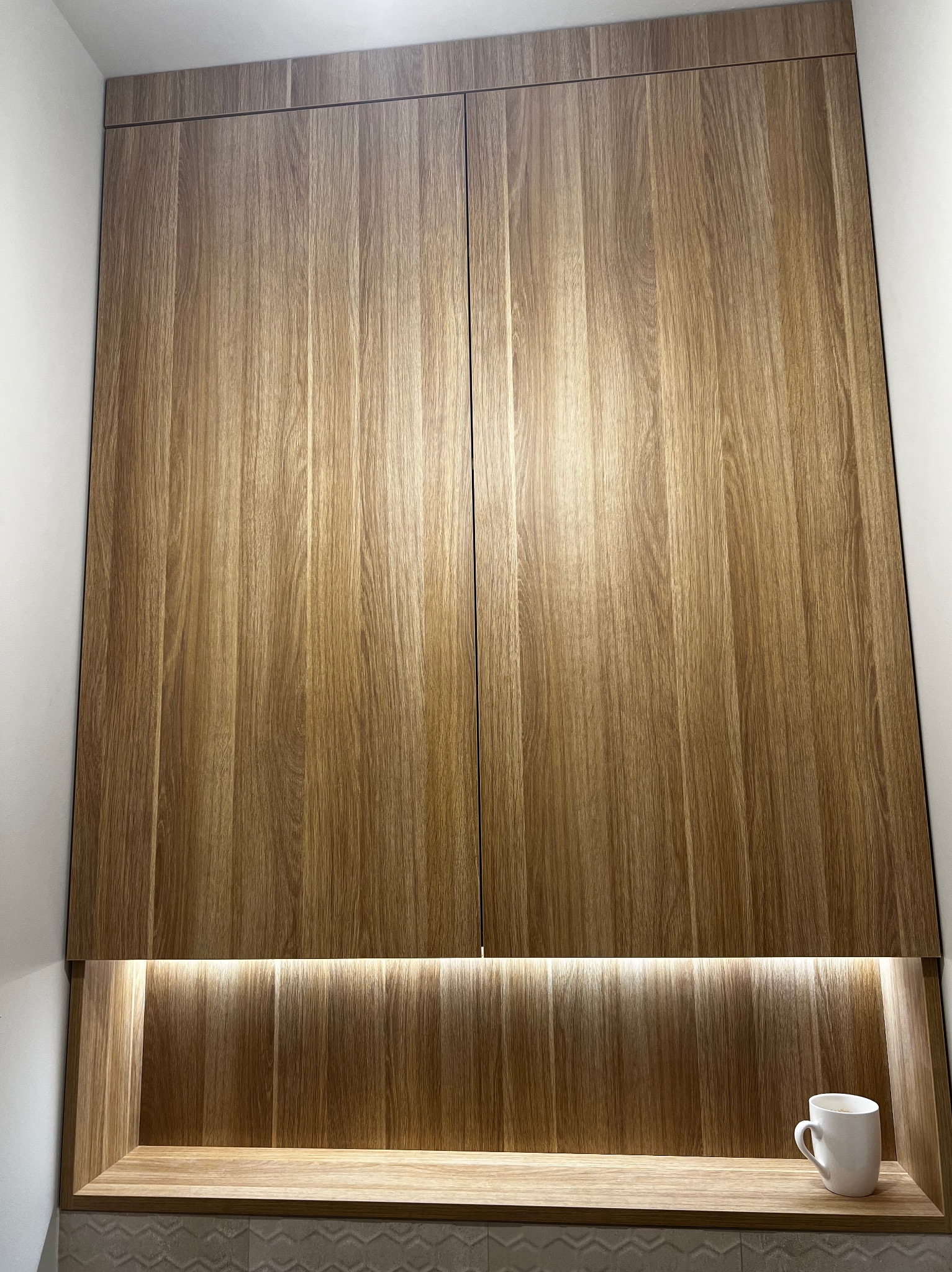 Drewniana szafka wisząca z podświetlaną wnęką, w której stoi biały kubek.