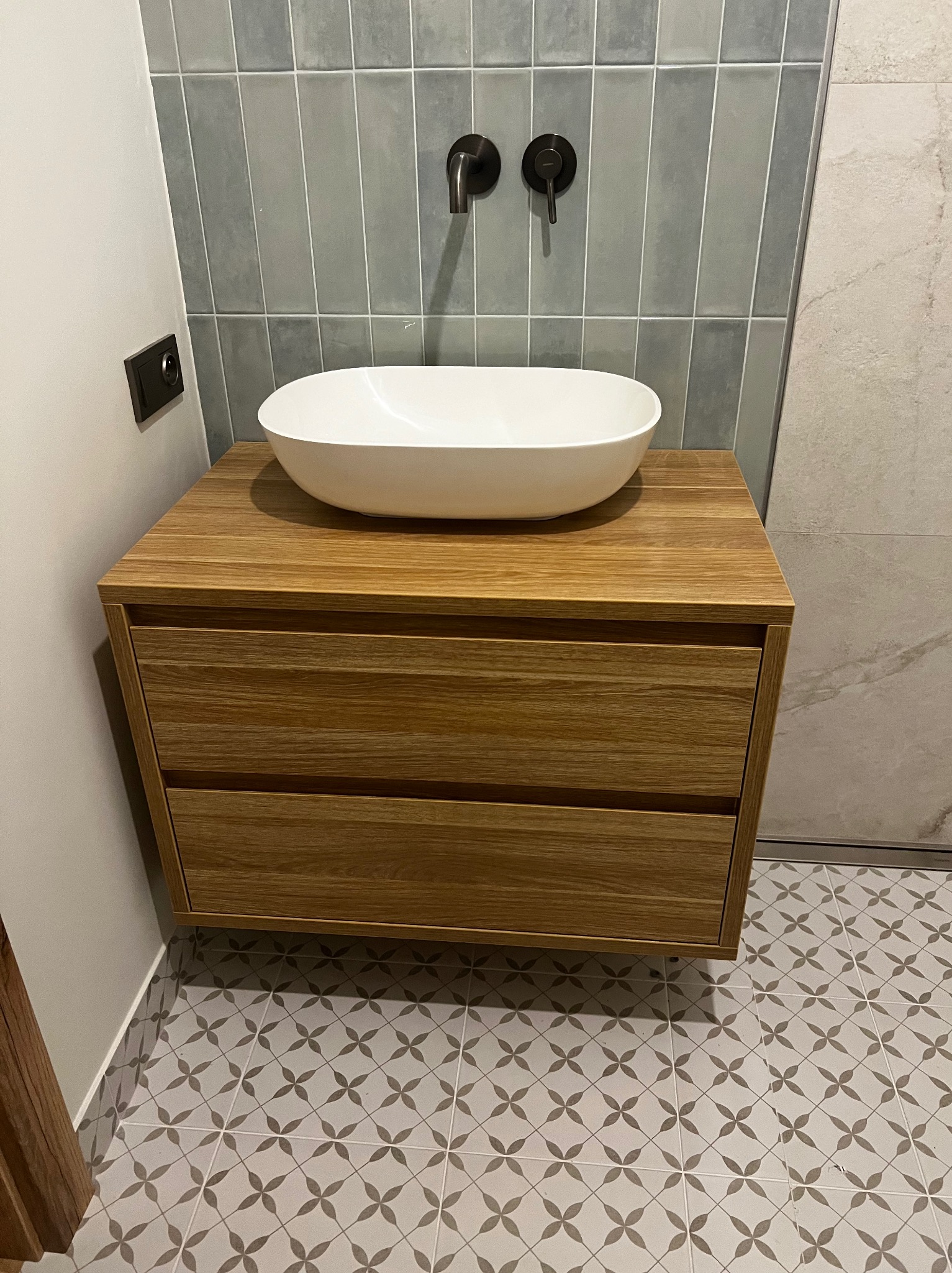 Łazienka z drewnianą szafką pod umywalkę z dwoma szufladami, białą owalną umywalką nablatową i grafitową armaturą na tle szarych płytek w pionowe pasy.