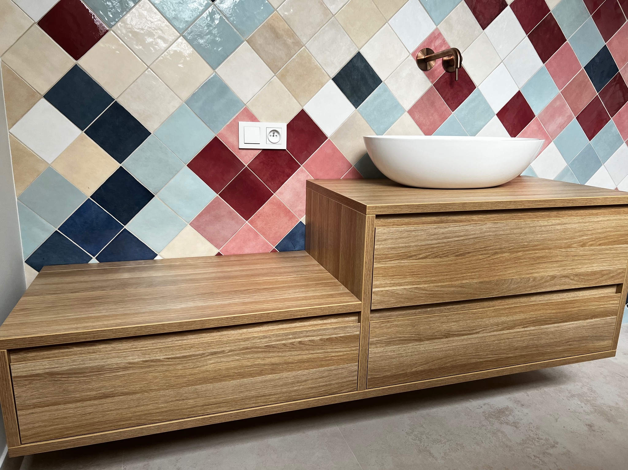 Drewniana szafka łazienkowa z dwiema szufladami i białą umywalką nablatową, na tle ściany z kolorowych, romboidalnych płytek ceramicznych i miedzianej baterii.