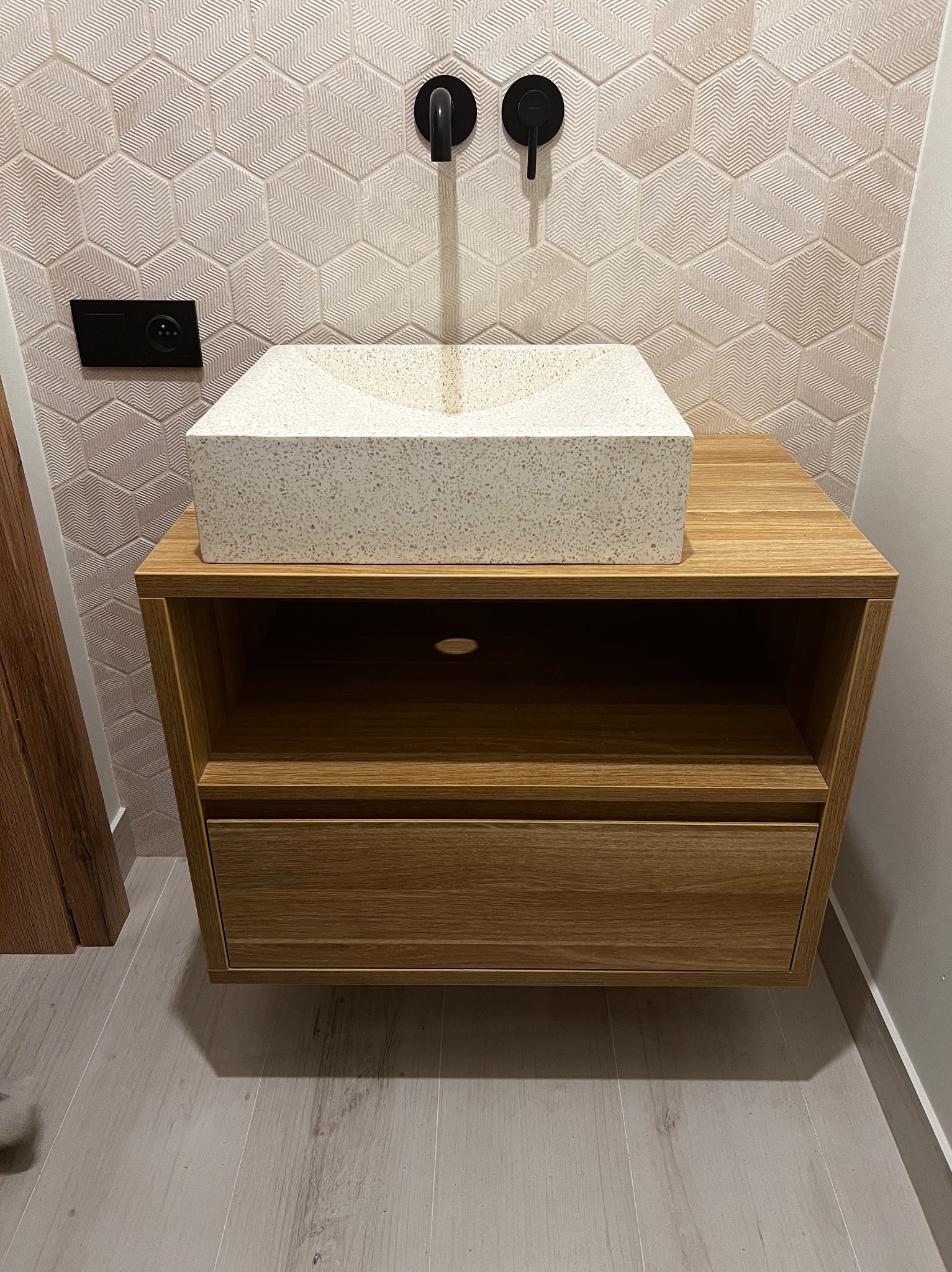 Łazienkowa szafka z drewna z szufladą i otwartą półką, na której stoi kwadratowa umywalka z jasnego lastryko, czarna bateria ścienna, tło z heksagonalnych płytek o subtelnym wzorze.
