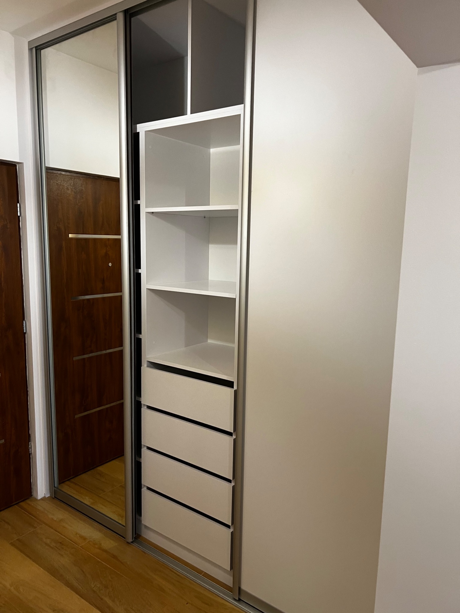 Nowoczesna szafa wnękowa z przesuwnymi drzwiami lustrzanymi i białymi półkami wewnątrz, widoczna w mieszkaniu w Warszawie. Na podłodze drewniane panele.