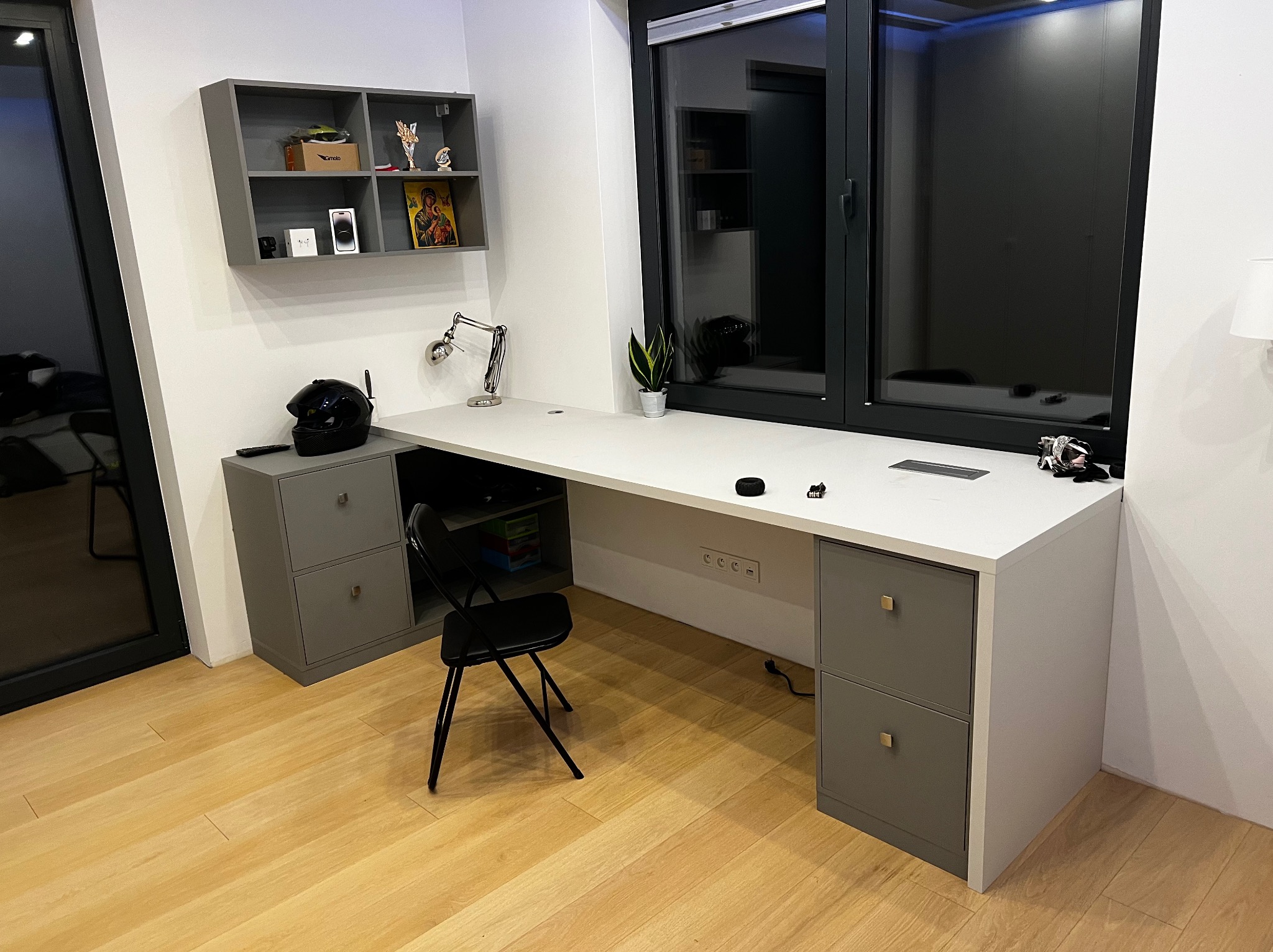 Nowoczesne biurko na wymiar z szarymi szafkami po bokach, biały blat, czarne krzesło składane, półka wisząca z dekoracjami, duże okno z czarną ramą, drewniana podłoga.