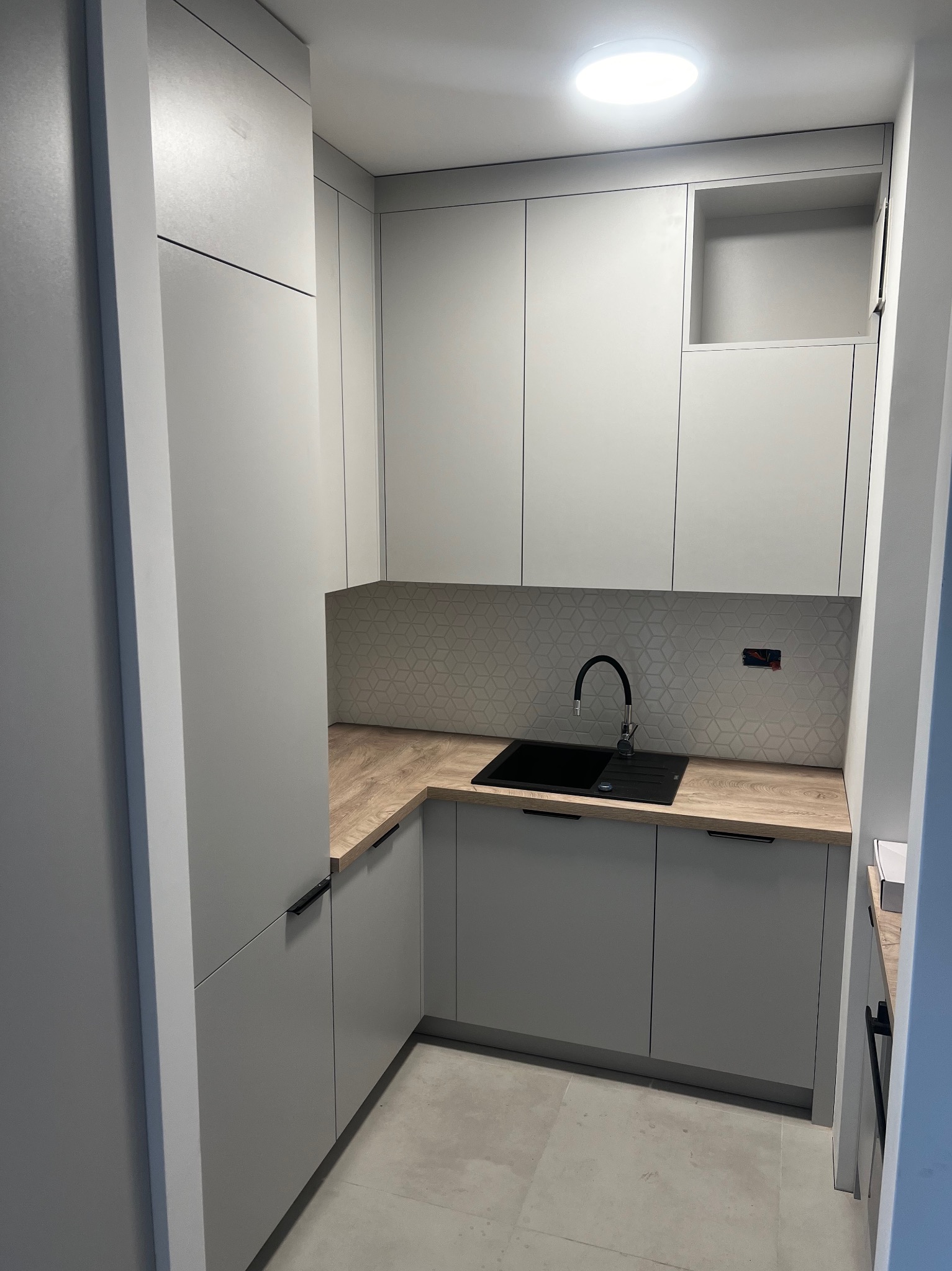 Jasnoszara zabudowa kuchenna z drewnianym blatem i czarnym zlewem, geometryczna mozaika na ścianie, widoczne gniazdko elektryczne bez osłonki.