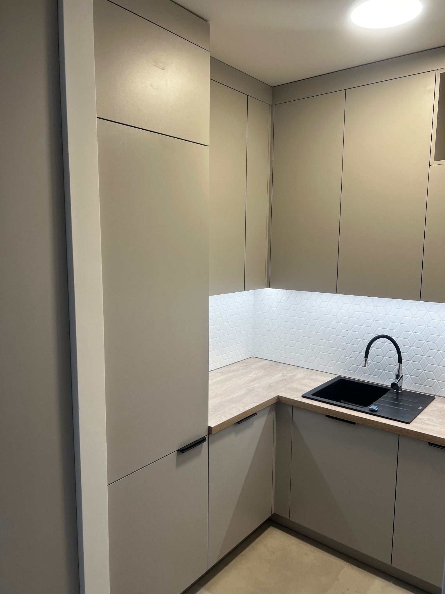 Beżowa zabudowa kuchenna z czarnym zlewem i baterią, heksagonalne płytki nad blatem o drewnianym wykończeniu, minimalistyczne uchwyty szafek.