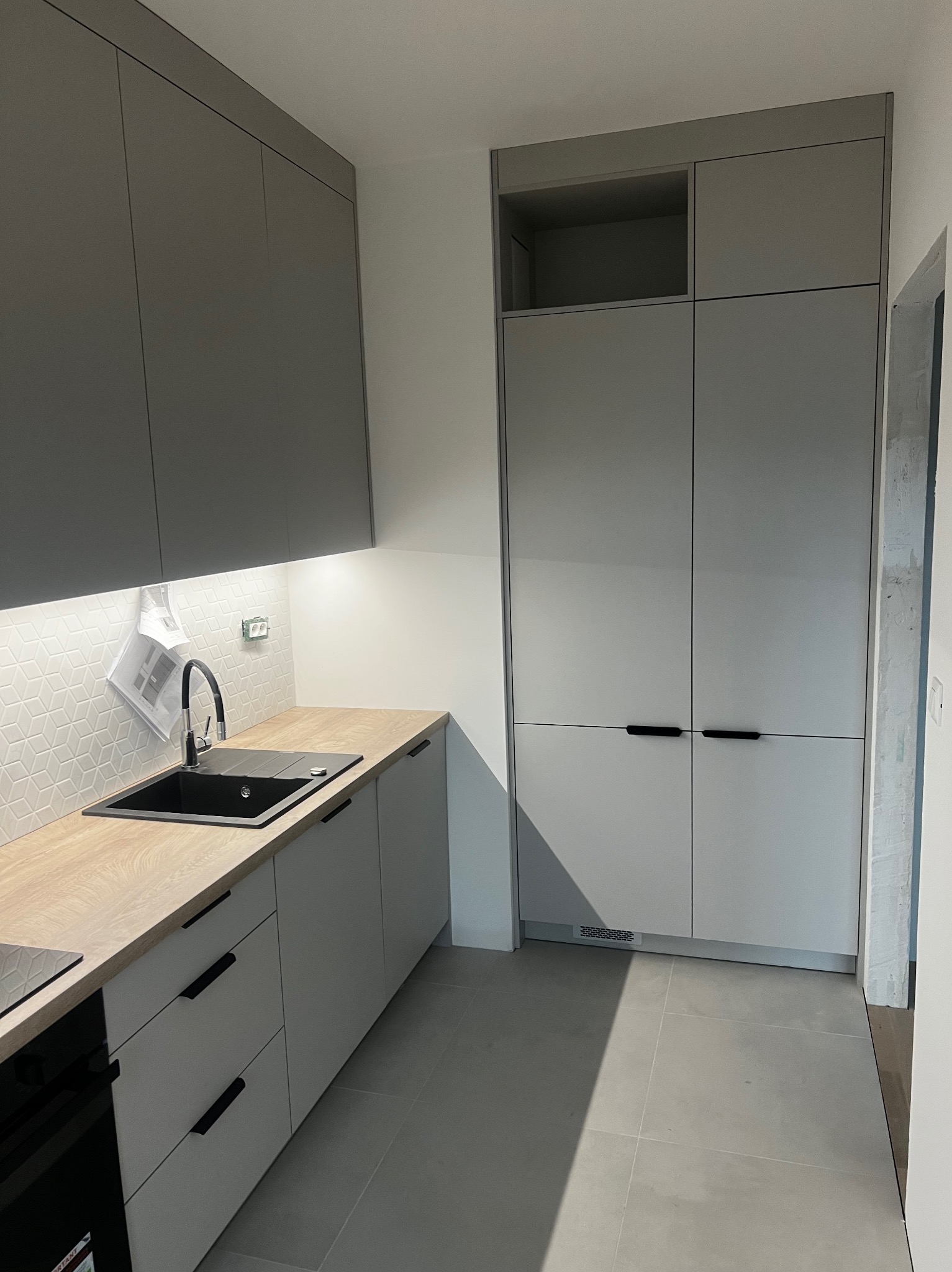 Szara kuchnia na wymiar z drewnianym blatem, czarnym zlewem i minimalistycznymi uchwytami, oświetlona ledami podszafkowymi, z wiszącymi dokumentami na ścianie.