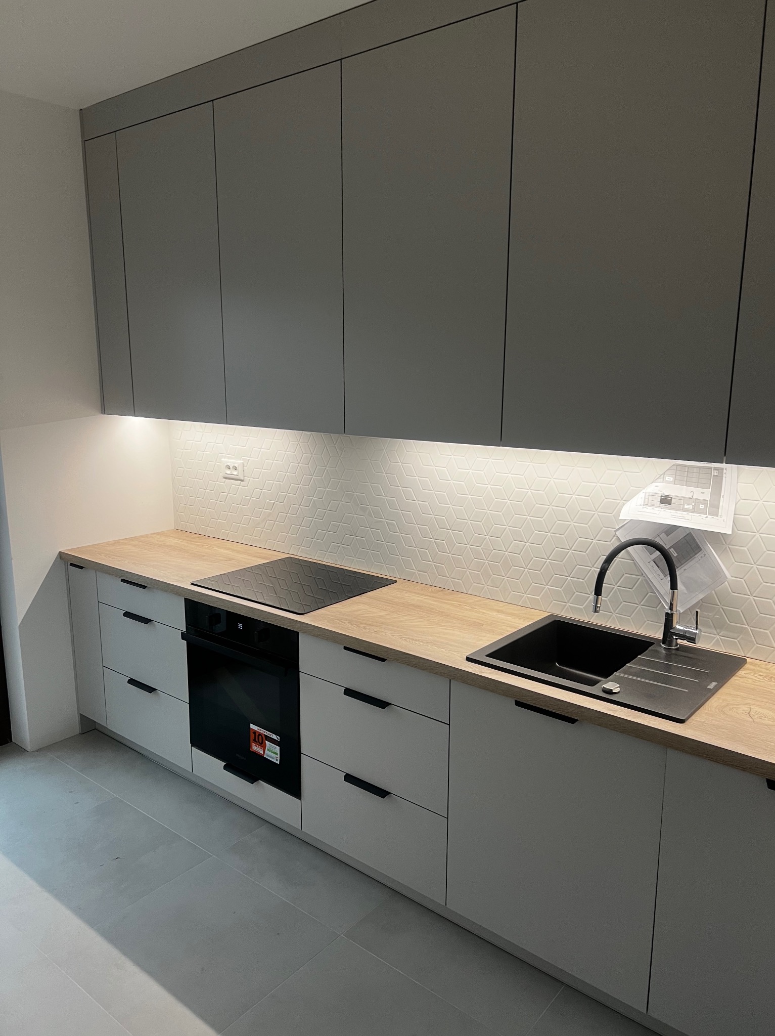 Szara zabudowa kuchenna z drewnianym blatem, czarnym zlewem i płytą indukcyjną, podświetlona listwą LED, z planami architektonicznymi przypiętymi do ściany.