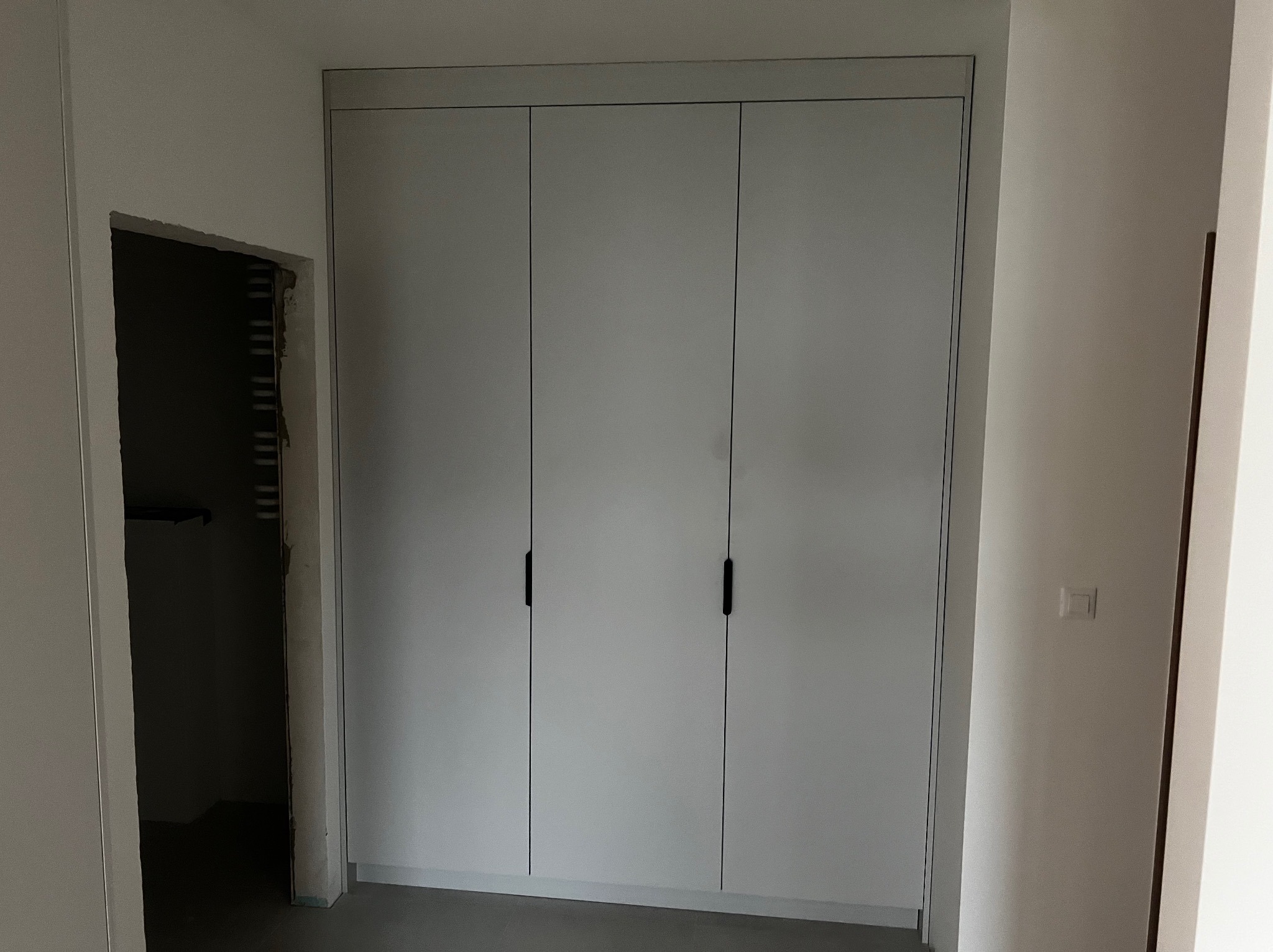 Trzydrzwiowa szafa wnękowa z szarymi frontami i czarnymi, pionowymi uchwytami, widoczna w trakcie montażu, z surowym otworem drzwiowym w tle.