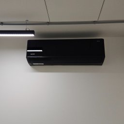 HZ KLIMA Henryk Zaworski - Czarna jednostka klimatyzacji wisząca na białej ścianie, widoczne szyny montażowe, lampa sufitowa i klapa rewizyjna.