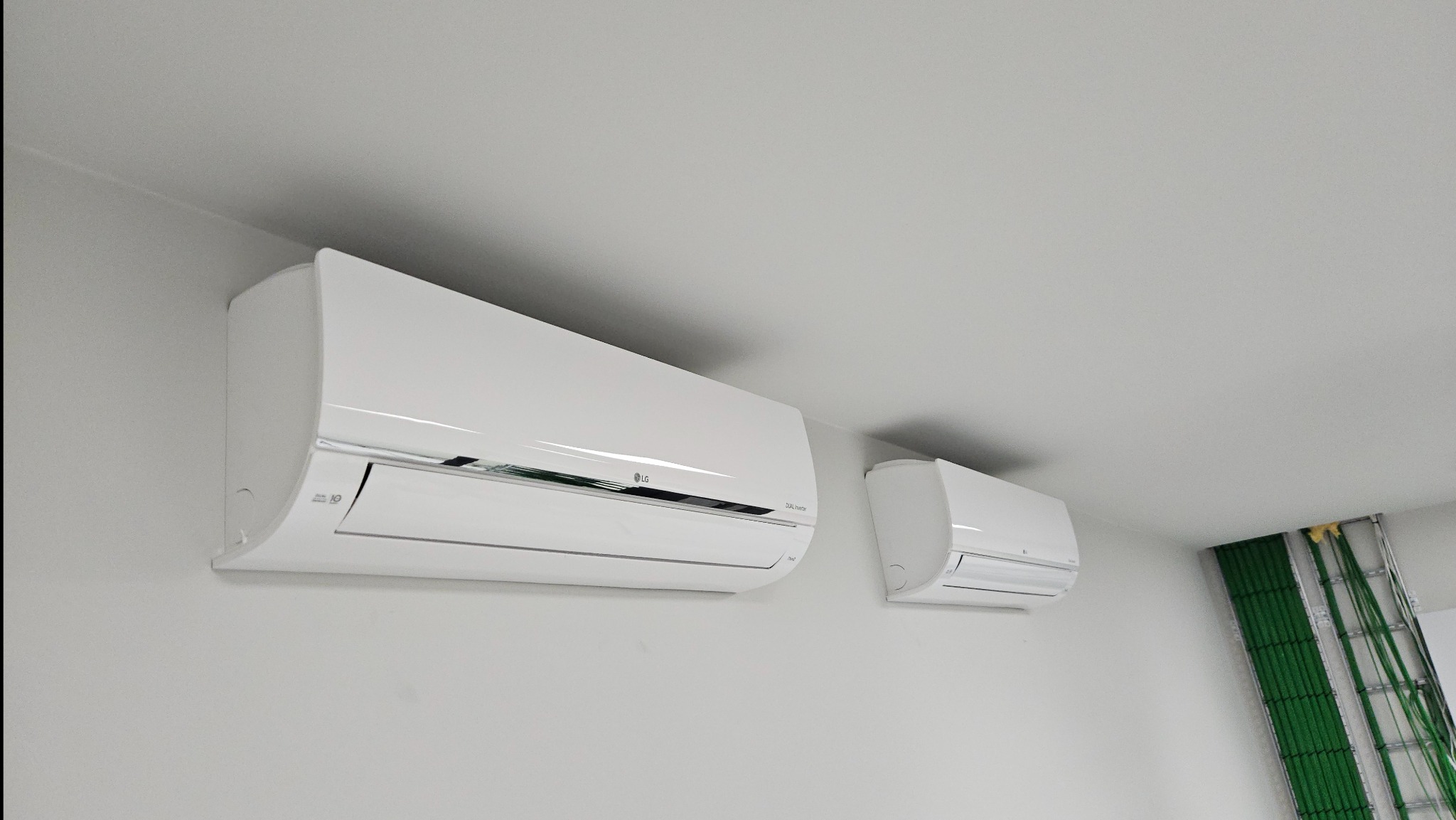 Dwa białe klimatyzatory ścienne LG o różnej wielkości, zamontowane w pomieszczeniu z białymi ścianami i sufitem, widoczne kable w korytku po prawej stronie.
