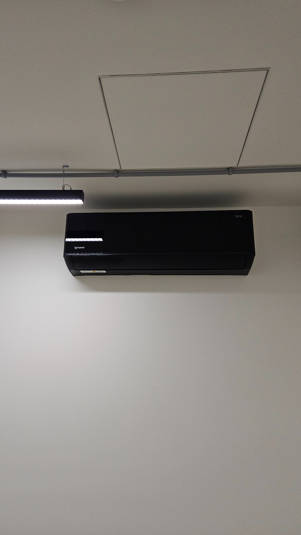 Czarna jednostka klimatyzacji wisząca na białej ścianie, widoczne szyny montażowe, lampa sufitowa i klapa rewizyjna.