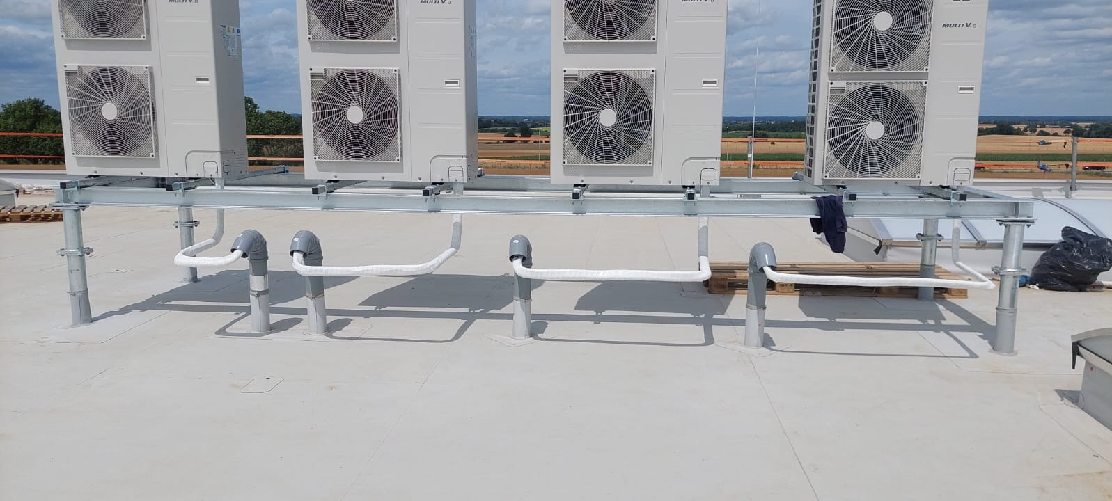 Instalacja klimatyzatorów LG Multi V na dachu budynku, widoczne jednostki zewnętrzne na metalowej konstrukcji wsporczej, rury odprowadzające skropliny z izolacją termiczną, w tle krajobraz.