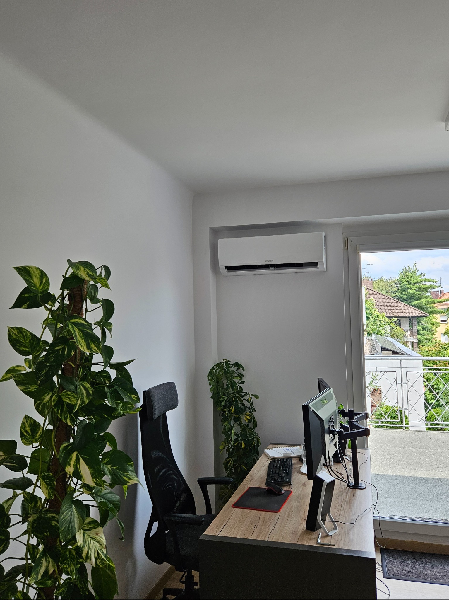 Biały klimatyzator na ścianie nad biurkiem w pokoju z balkonem, widoczne rośliny doniczkowe i sprzęt komputerowy.