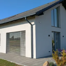 Nowoczesny dom jednorodzinny z jasną elewacją, szarym dachem i dużymi oknami z żaluzjami, widok z zewnątrz na taras z meblami ogrodowymi.