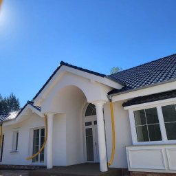 DomKraft Anton Klimenka - Elewacja świeżo otynkowanego, parterowego domu z ciemną dachówką, widoczne żółte rury spustowe i fragment elewacji w budowie.