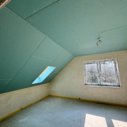 DomKraft Anton Klimenka - Poddasze w trakcie prac wykończeniowych: zielone płyty gipsowo-kartonowe na skosach sufitu, okno dachowe i okno pionowe z widokiem na drzewa, surowe ściany przygotowane do malowania, żółte...