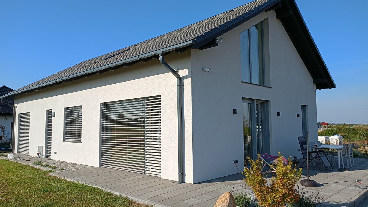 Nowoczesny dom jednorodzinny z jasną elewacją, szarym dachem i dużymi oknami z żaluzjami, widok z zewnątrz na taras z meblami ogrodowymi.