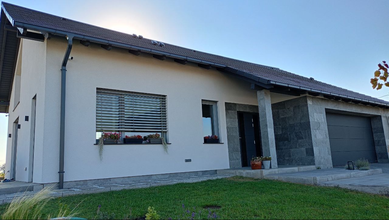 Nowoczesny dom jednorodzinny z białymi ścianami, ciemną dachówką i szarymi elementami wykończeniowymi, kwiaty w donicach pod oknami, widok z zewnątrz na tle błękitnego nieba.