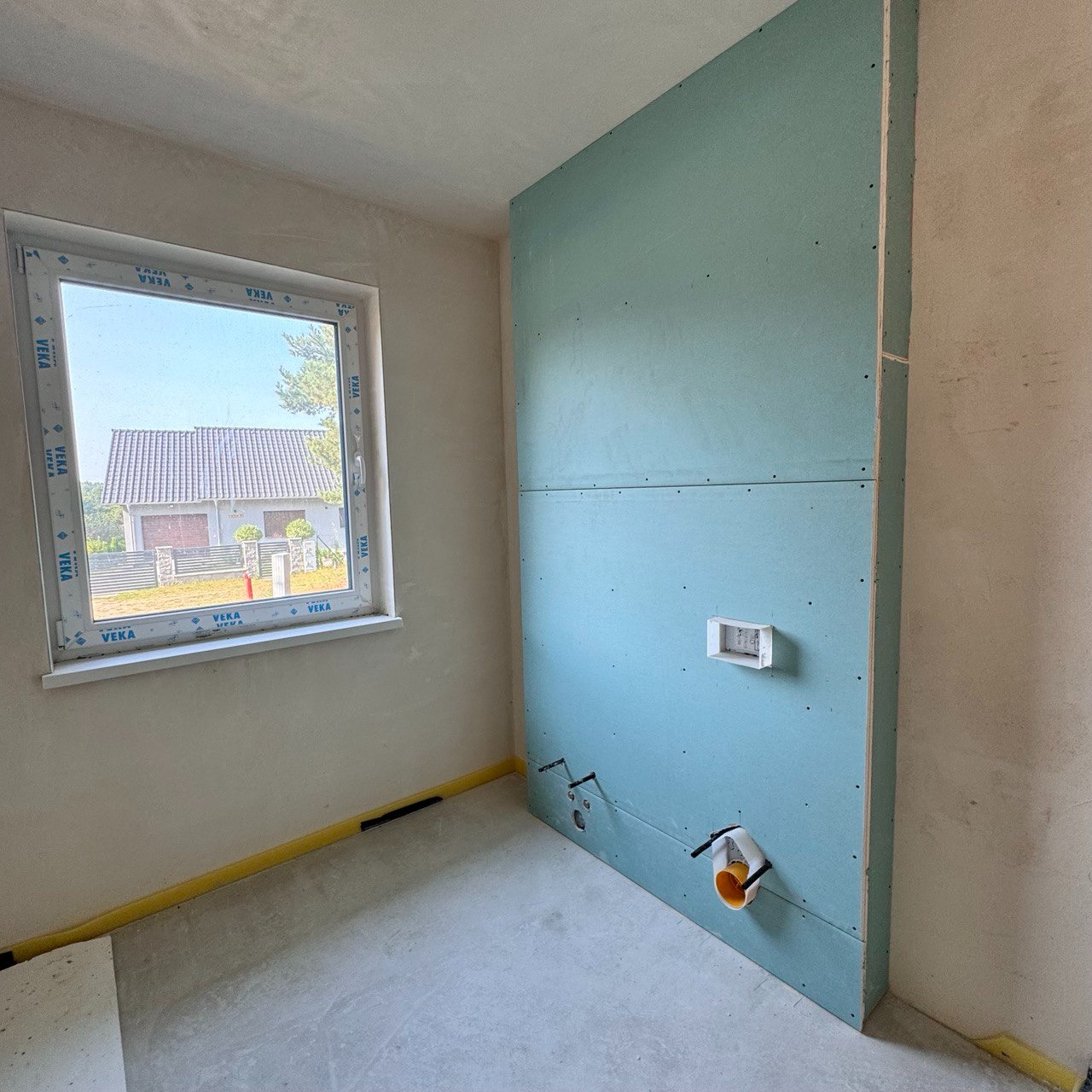 Montaż płyty karton gips na ścianie w łazience z widocznymi instalacjami hydraulicznymi i stelażem podtynkowym, okno z widokiem na dom.