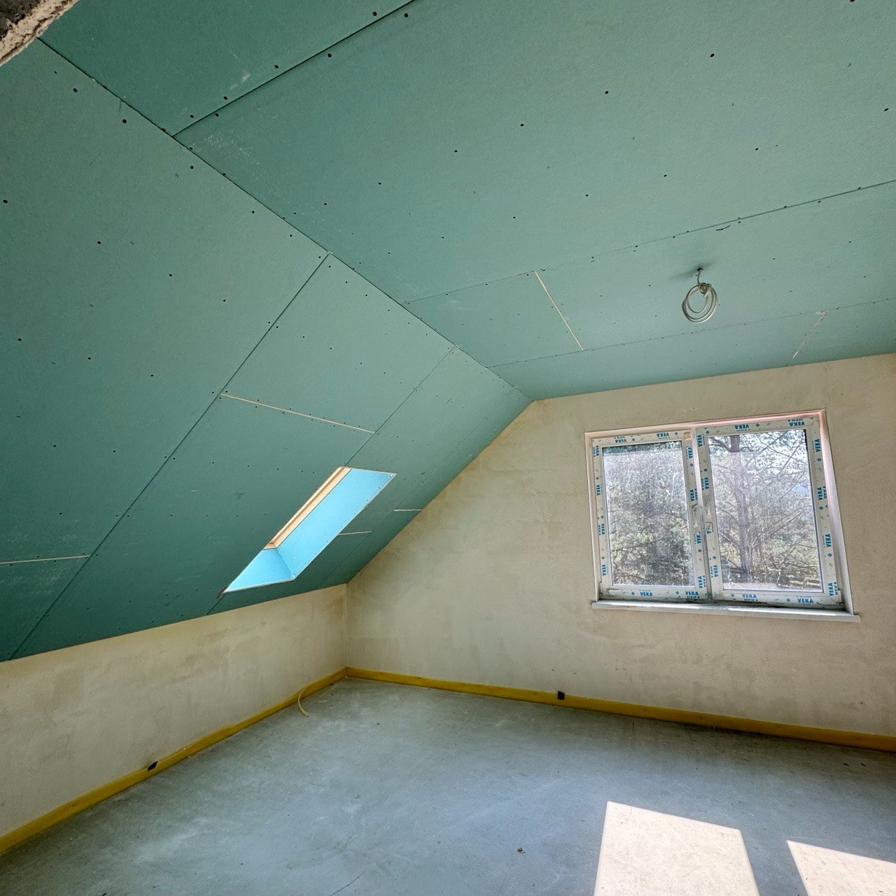 Poddasze w trakcie prac wykończeniowych: zielone płyty gipsowo-kartonowe na skosach sufitu, okno dachowe i okno pionowe z widokiem na drzewa, surowe ściany przygotowane do malowania, żółte...