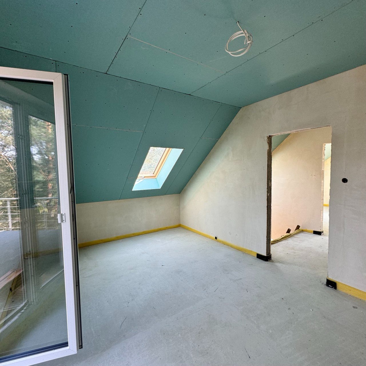 Pokój na poddaszu w Gorzowie Wielkopolskim w trakcie prac wykończeniowych: sufit pokryty zieloną płytą, okno dachowe, ściany w stanie surowym, widoczne taśmy zabezpieczające podłogę.