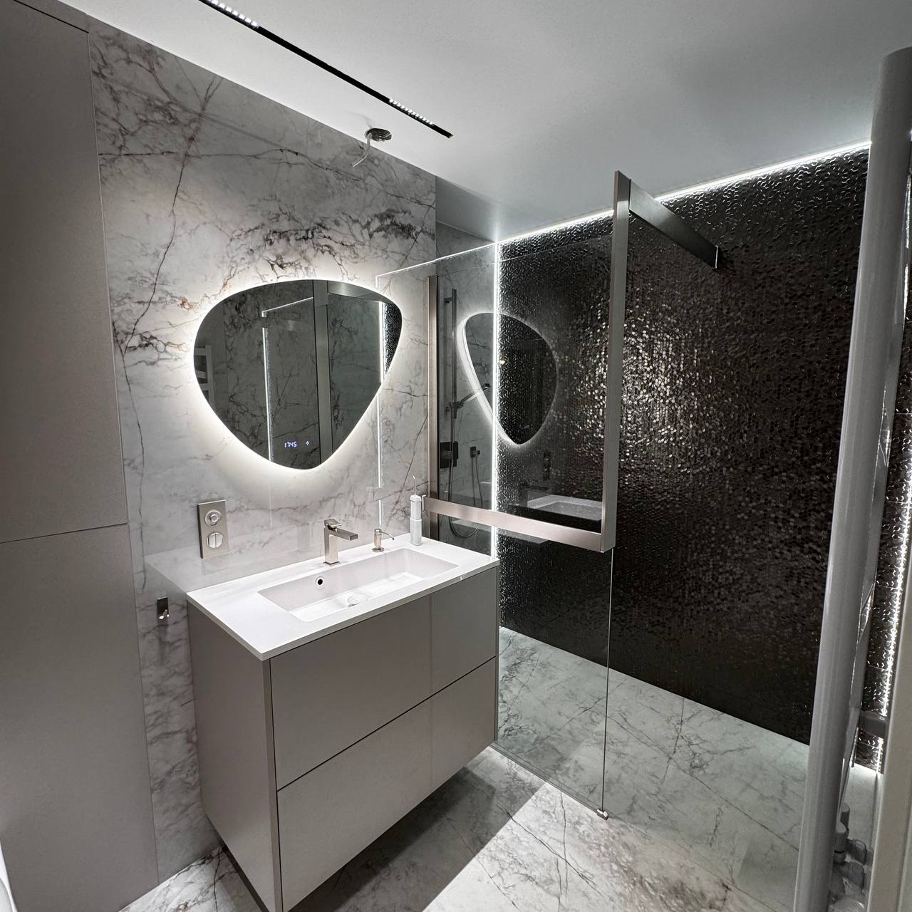 Nowoczesna łazienka z marmurowymi ścianami, podświetlanym lustrem o nieregularnym kształcie i szklaną kabiną prysznicową. Minimalistyczny design, szaro-biała kolorystyka.