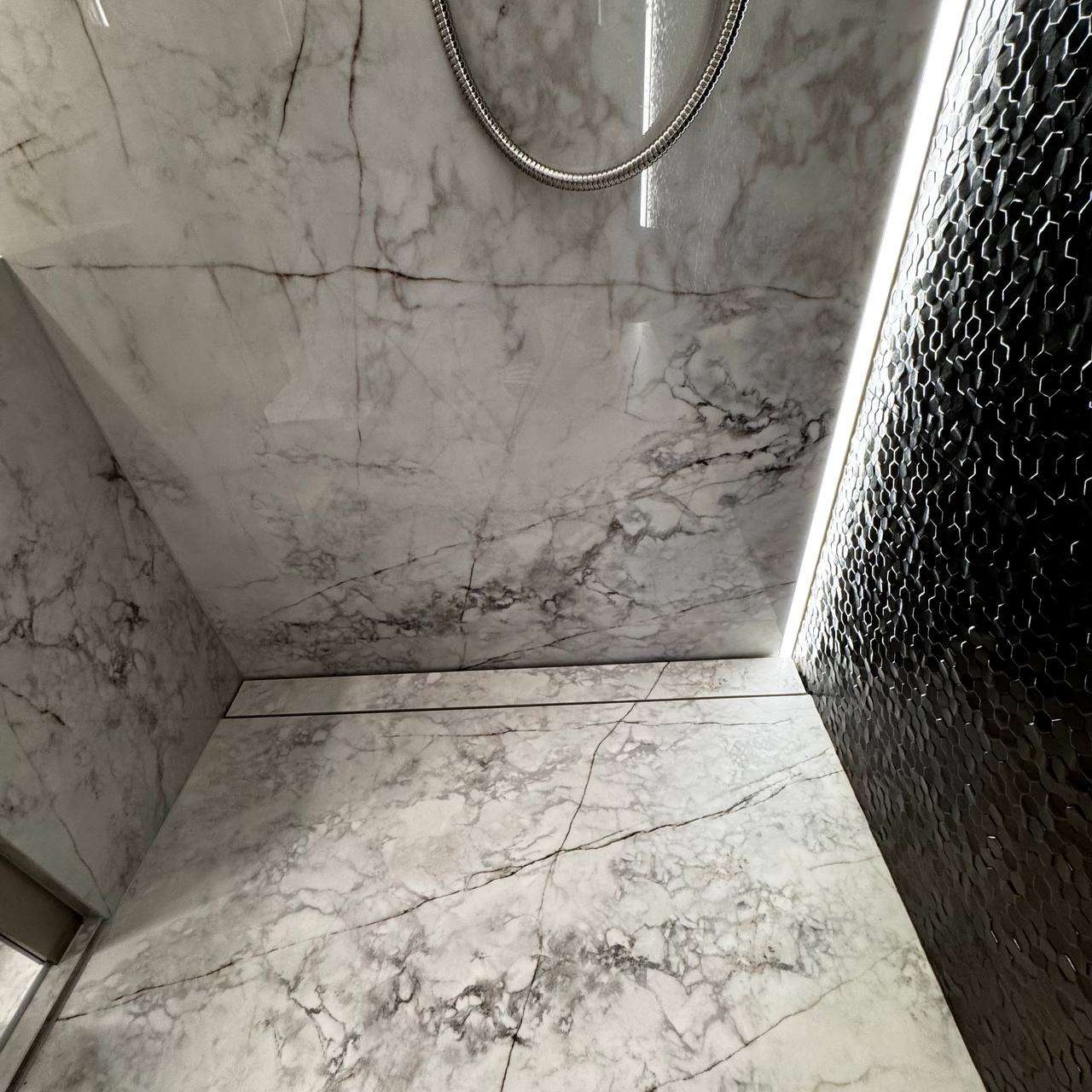 Wykończona kabina prysznicowa z białymi płytami imitującymi marmur i czarną mozaiką heksagonalną. Widoczna słuchawka prysznicowa i oświetlenie LED.