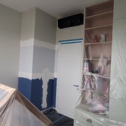 Robert Kelm - Pokój w trakcie przygotowań do odświeżenia ścian: zabezpieczone folią meble, fragment ściany z widocznymi pasami różnych kolorów przed nałożeniem jednolitej warstwy farby.