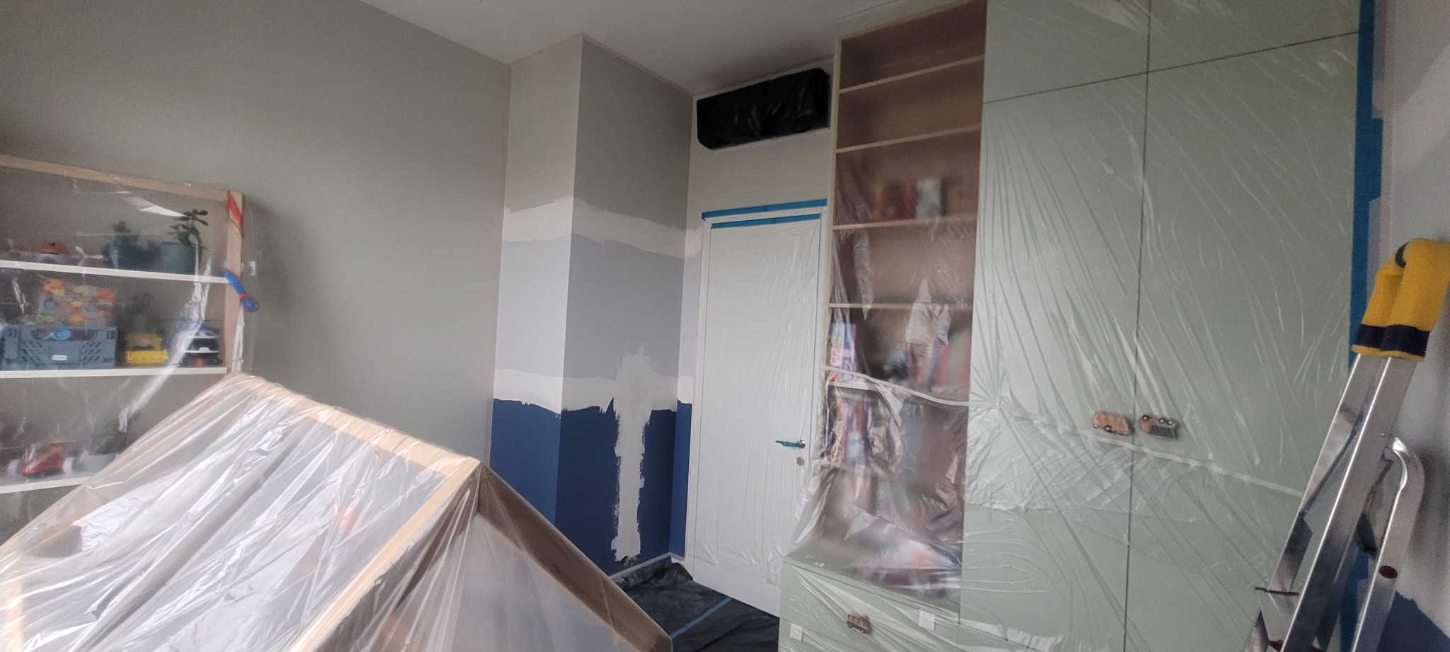 Pokój w trakcie przygotowań do odświeżenia ścian: zabezpieczone folią meble, fragment ściany z widocznymi pasami różnych kolorów przed nałożeniem jednolitej warstwy farby.