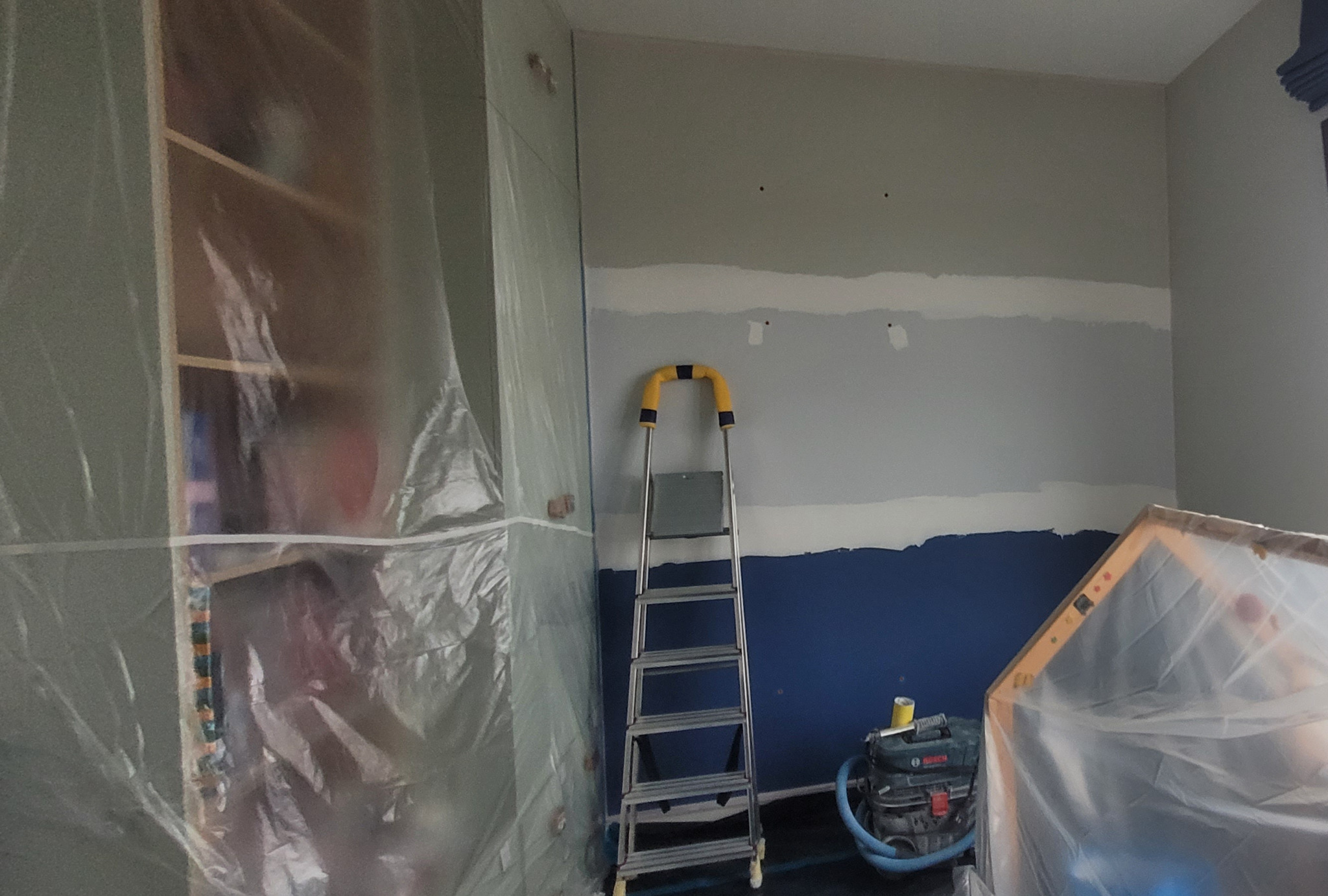 Przygotowanie ściany do malowania: zabezpieczone folią meble, ściana z fragmentami w kolorze niebieskim i szpachlowanymi pasami, drabina aluminiowa z żółtymi elementami, odkurzacz przemysłowy w tle.
