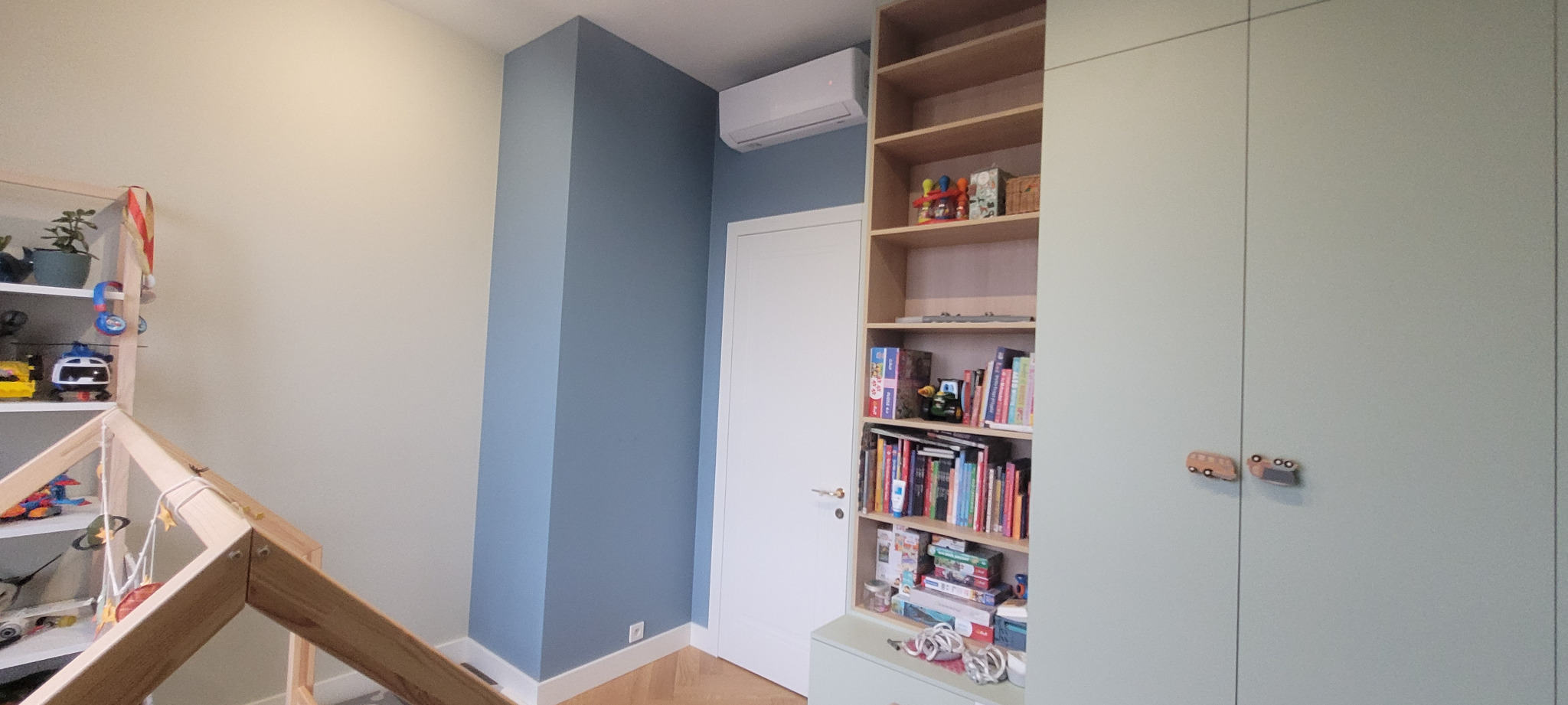 Pokój dziecięcy po remoncie: ściana pomalowana na dwa kolory (kremowy i niebieski), białe drzwi, klimatyzator, zabudowa meblowa z szafą i regałem na książki, drewniana podłoga w jodełkę.