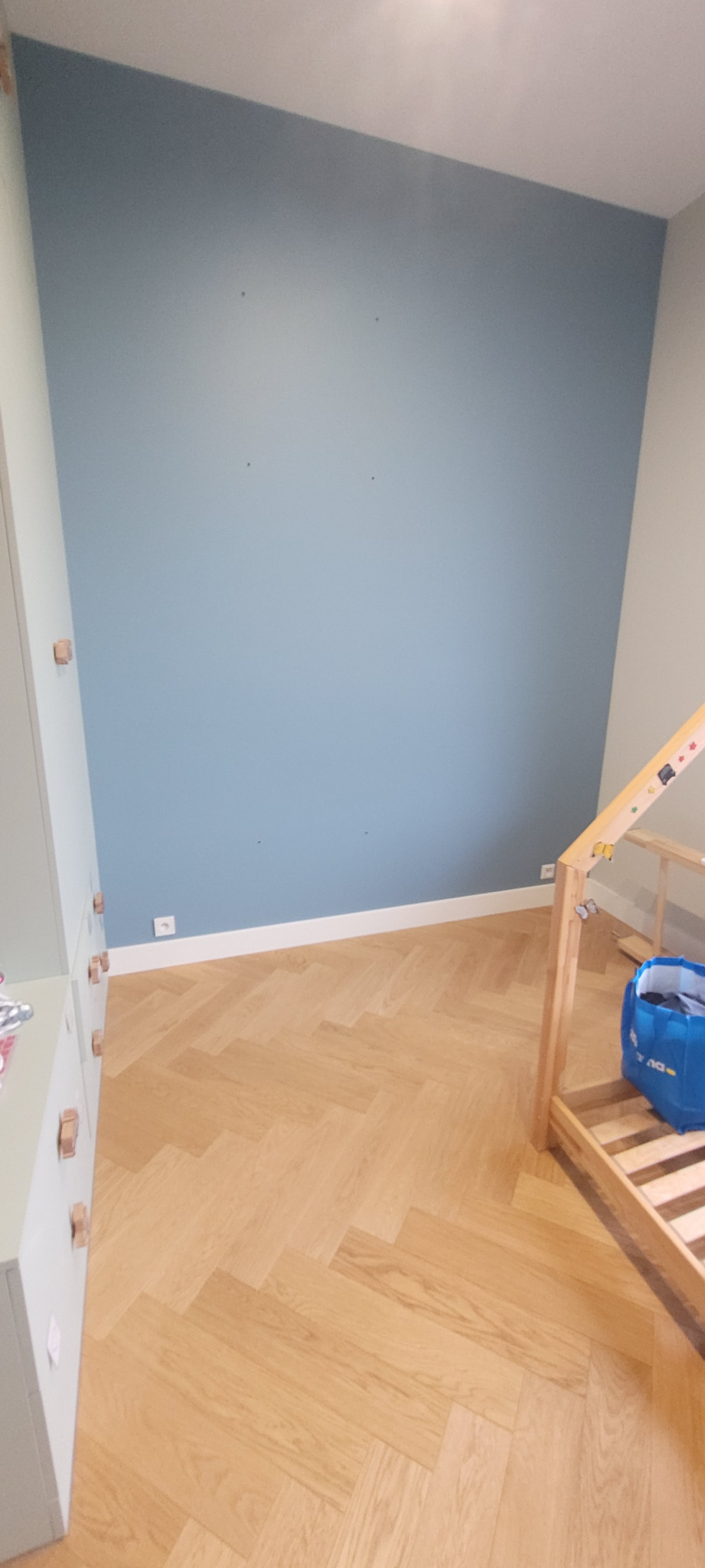 Świeżo pomalowana ściana w odcieniach błękitu w pokoju dziecięcym, widoczne ślady po kołkach, drewniana podłoga ułożona w jodełkę, biała listwa przypodłogowa i fragment szafy z drewnianymi uchwytami.